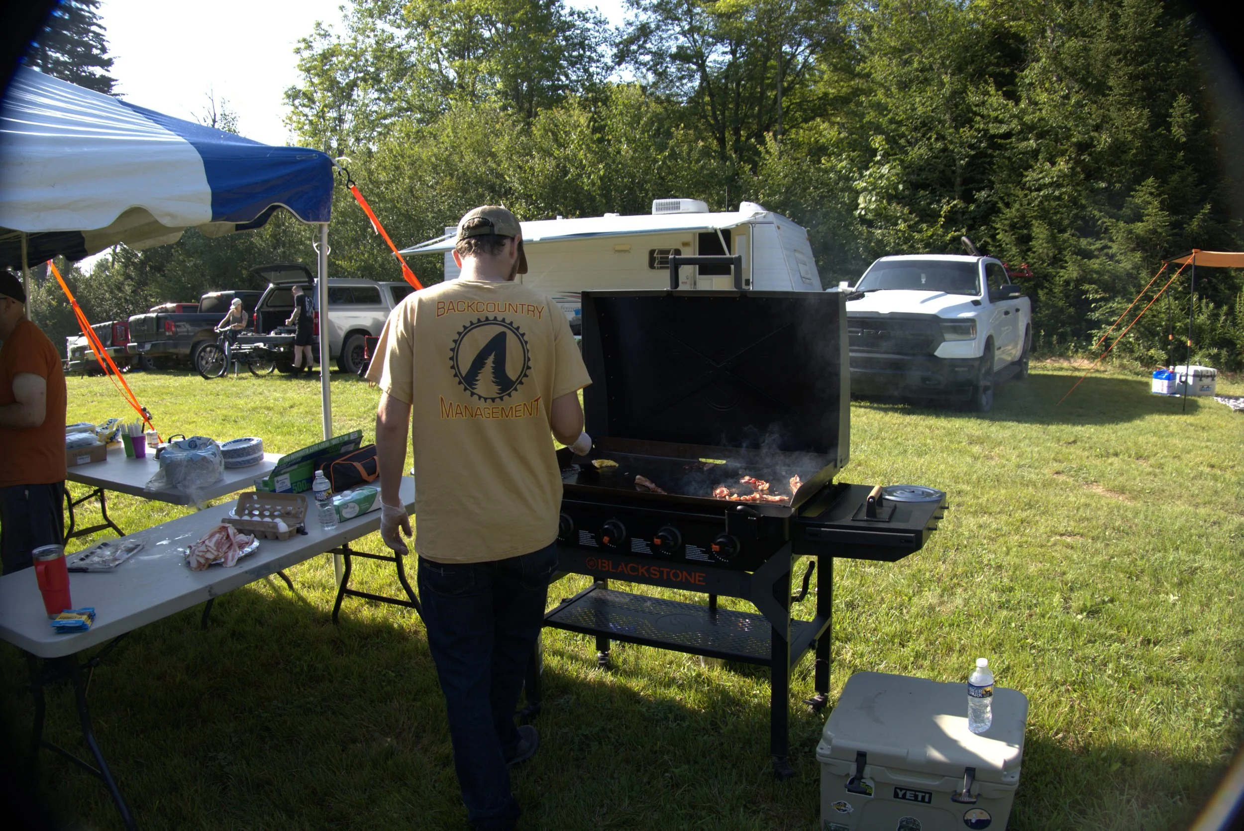 Person grilling meat on a barbecue grill outdoors with camping equipment, trucks, and trees in the background.