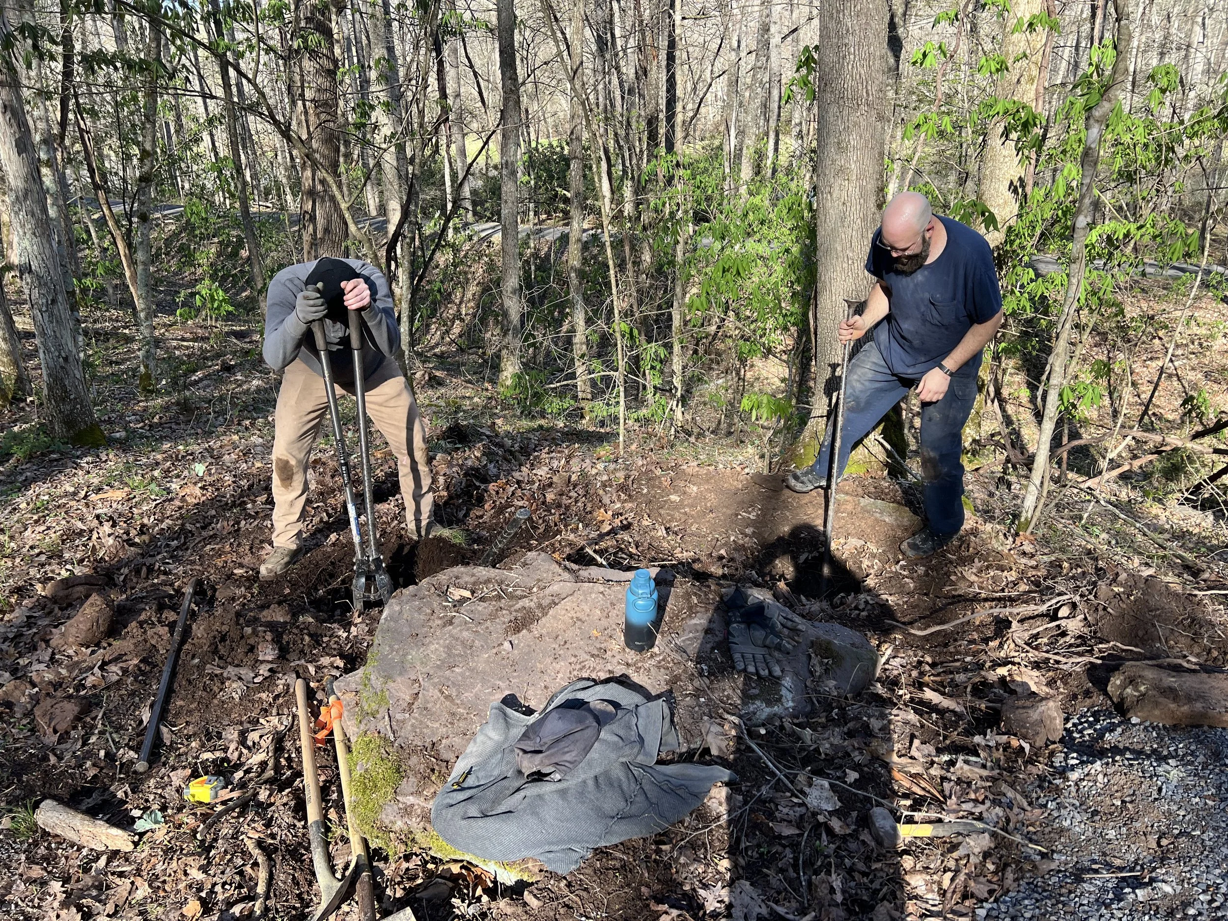 Two men digging holes in a forest, surrounded by trees and fallen leaves, with tools and a gray backpack on the ground.