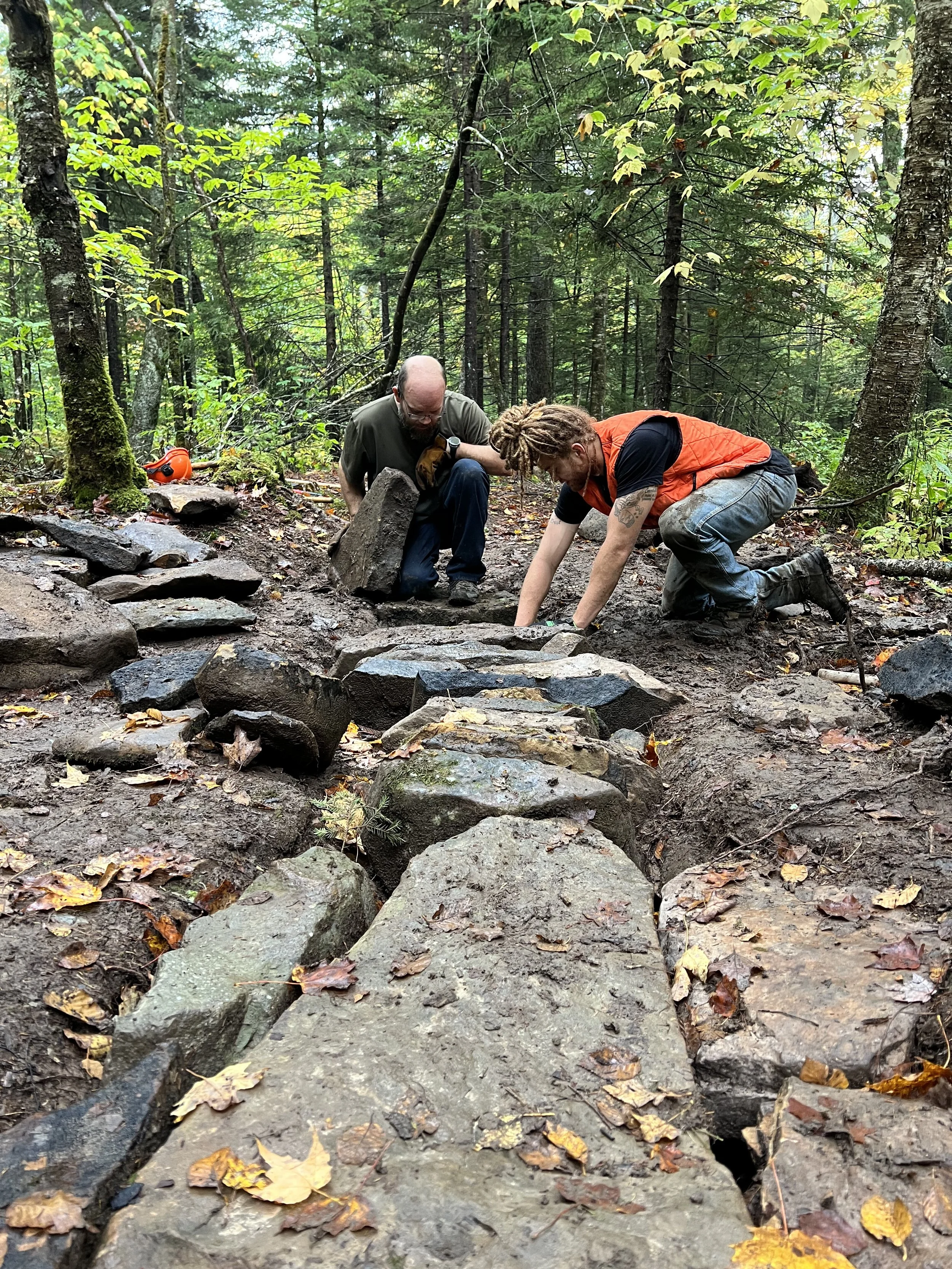 Two men working on a stone pathway in a forest, one kneeling and the other crouching, surrounded by trees and fallen leaves.