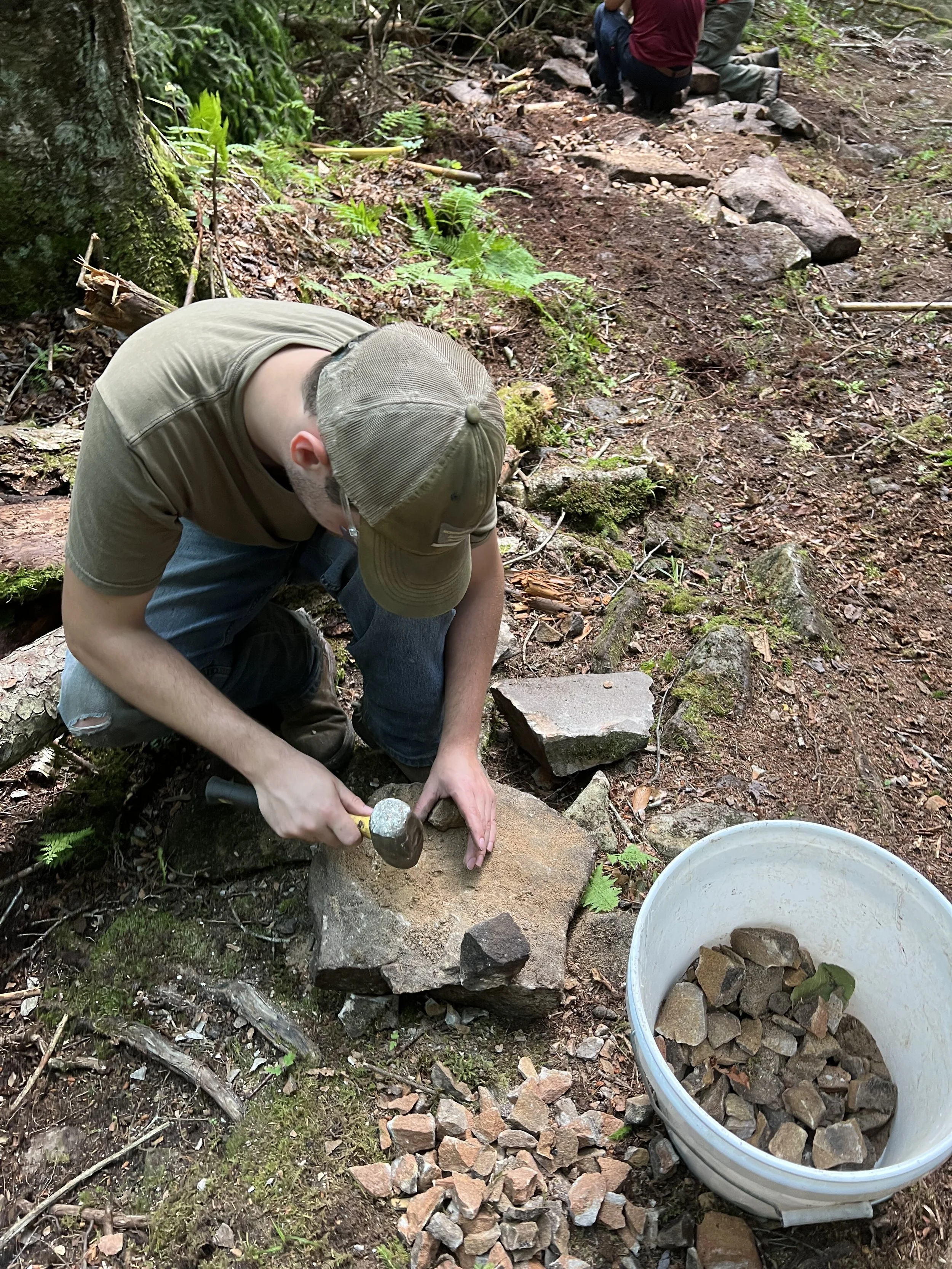 A person crouching down and using a hammer and chisel to split a large rock in a forest, with a bucket of small rocks nearby.
