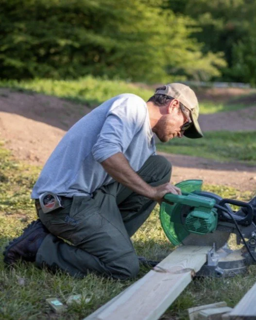 A man wearing a cap and sunglasses is kneeling outdoors, operating a green circular saw on a wooden plank.