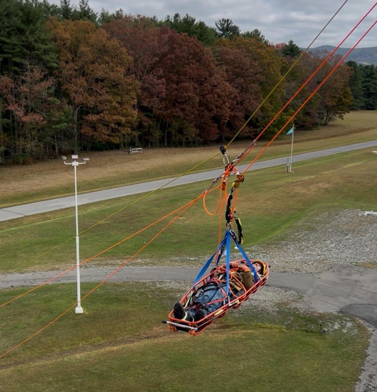 A rescue stretcher with gear hanging from a cable system high above a grassy area, used for aerial evacuation during outdoor activities or emergencies.