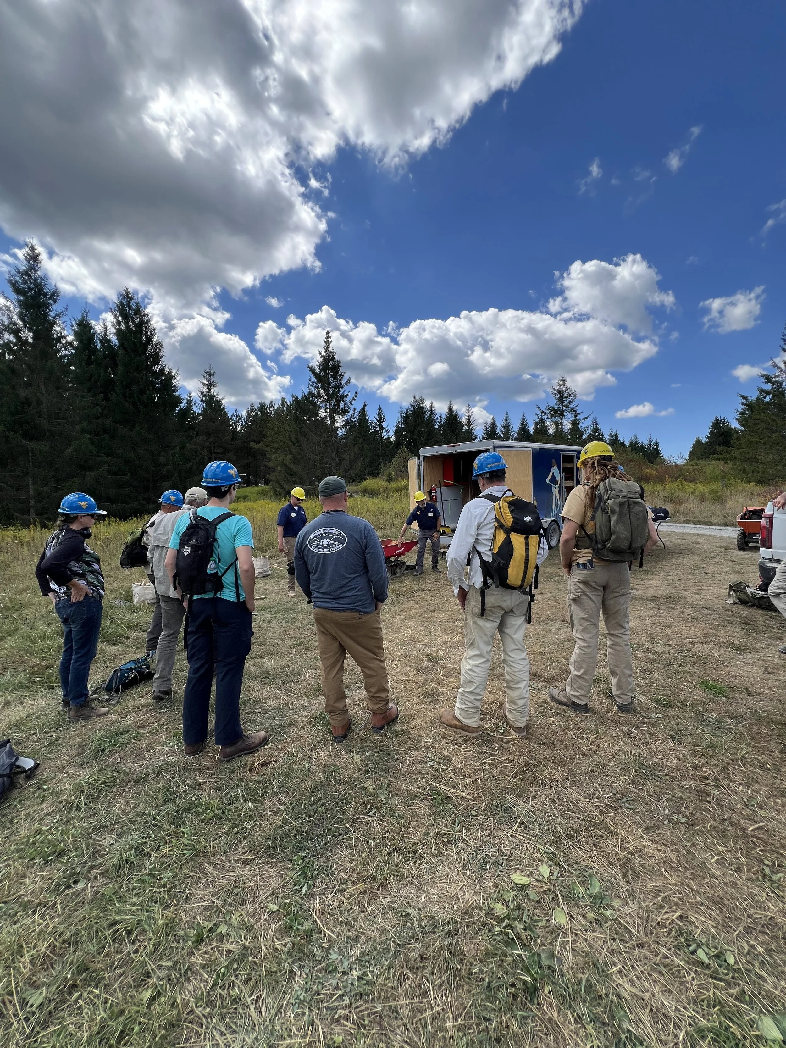 Group of people wearing safety helmets gathered outdoors on a grassy field with trees and a blue sky with clouds, attending a training or briefing near an enclosed trailer.