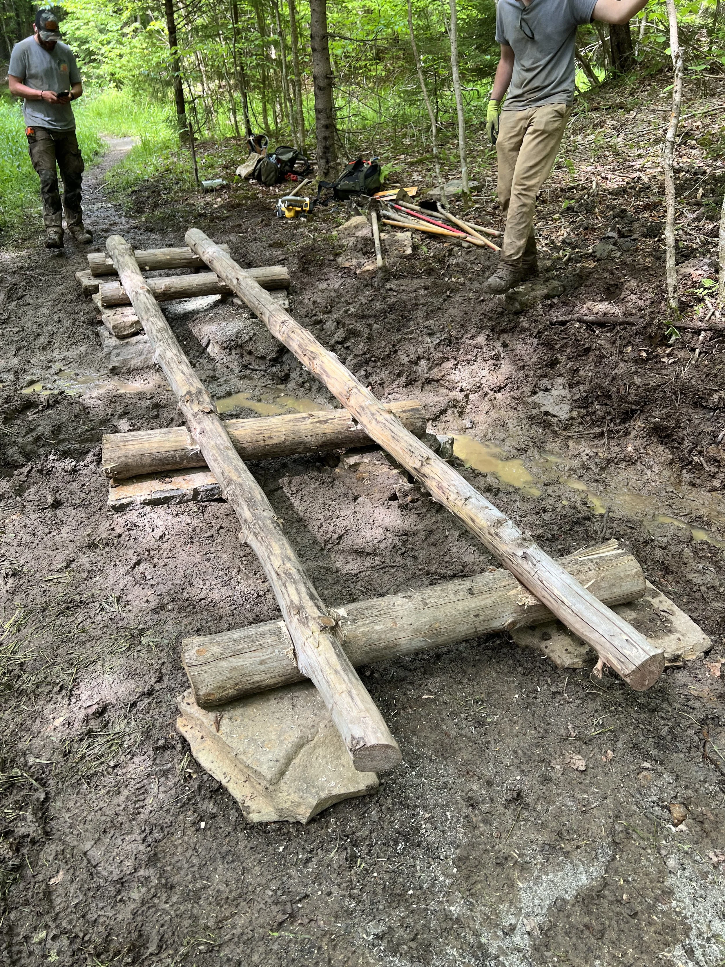 Two men working on constructing a wooden bridge over muddy terrain in a forest, with tools, bags, and equipment nearby.