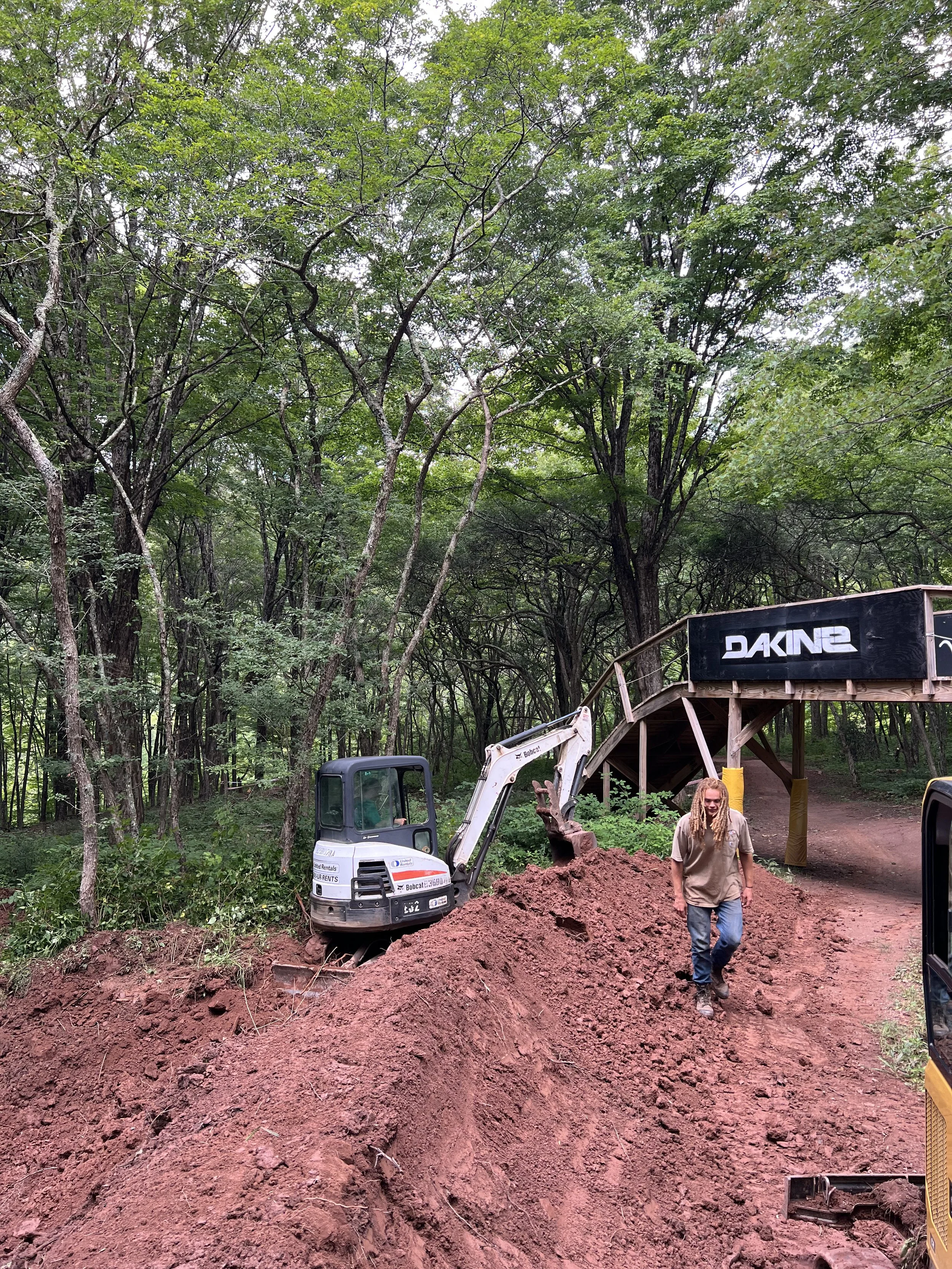 A person with long dreadlocks walking on a dirt path beside an excavator in a wooded area, with a bridge labeled 'DAKINE' in the background.