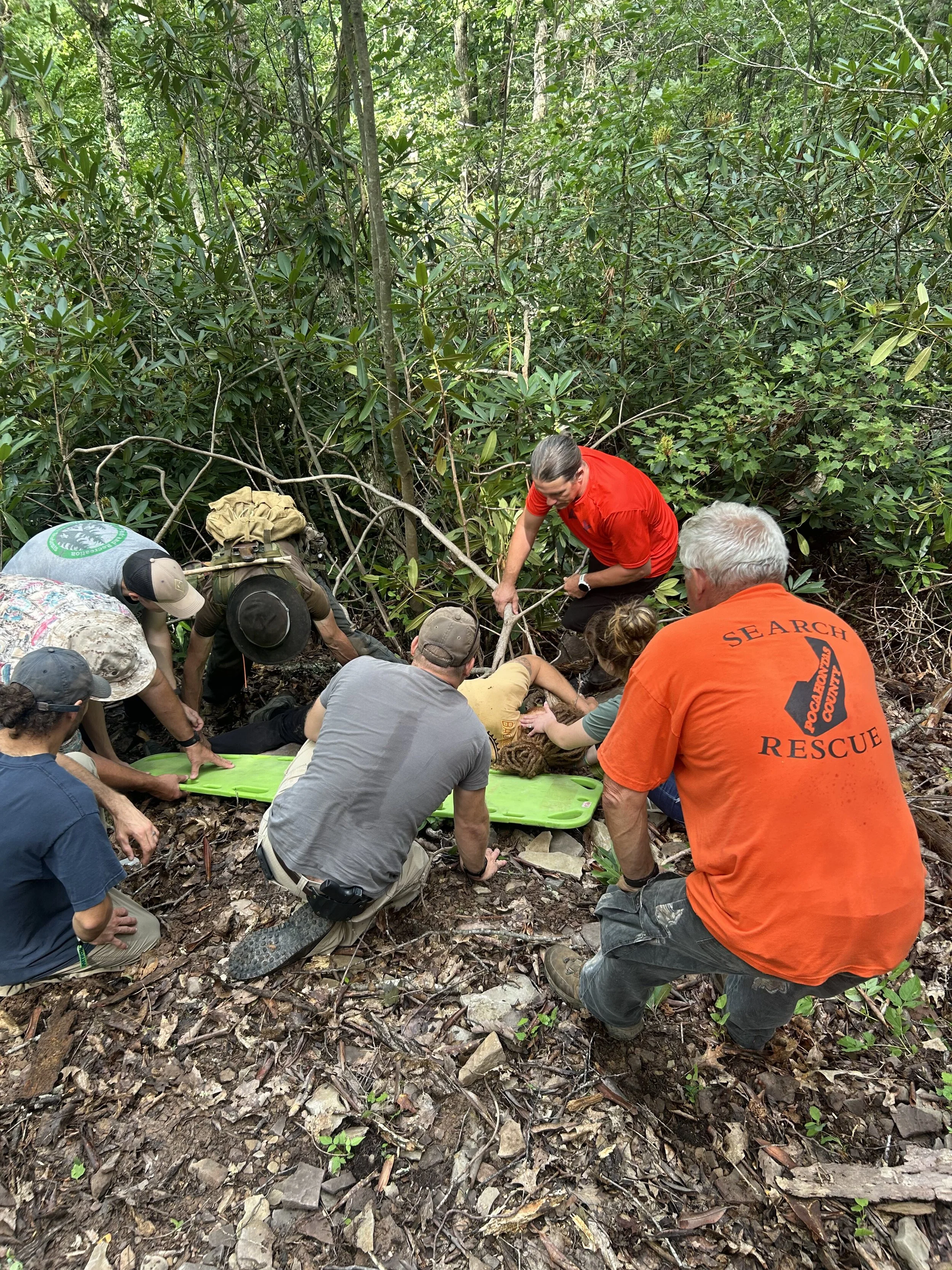 A group of rescue workers assisting a person in a wooded area with dense greenery. The team is focused on the person lying on a green rescue board, with some members holding and supporting them while others prepare equipment.