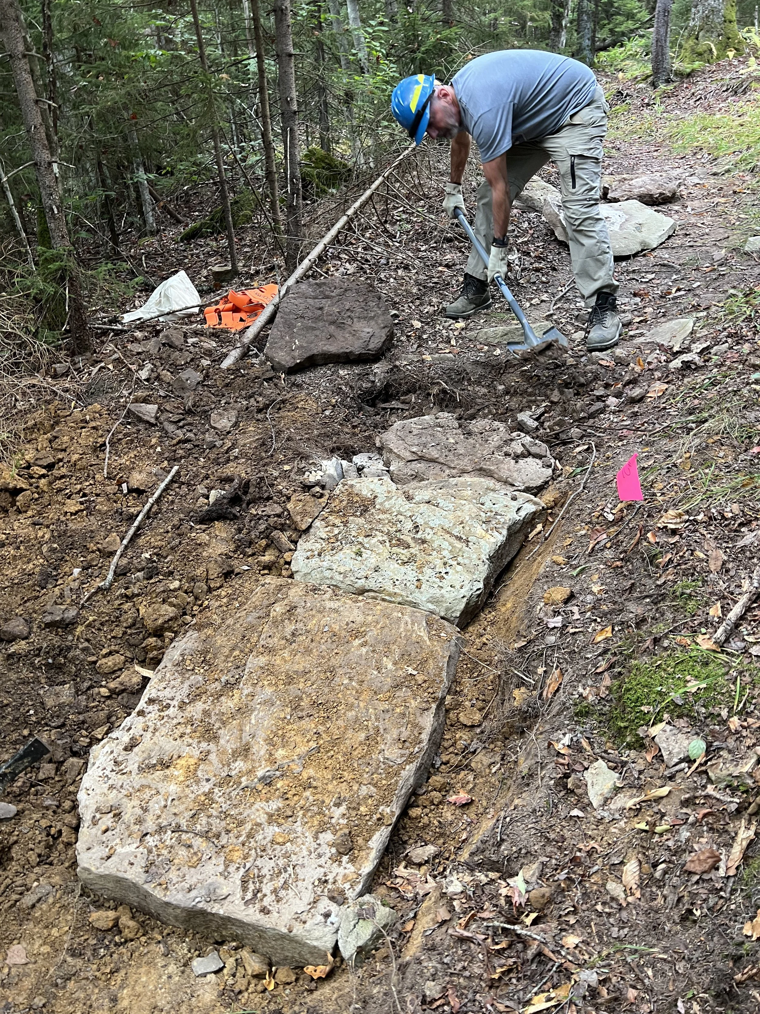 A person wearing a blue helmet, gray t-shirt, and cargo pants is working on a trail in a wooded area, using a hammer or similar tool to move or adjust large rocks along the trail.