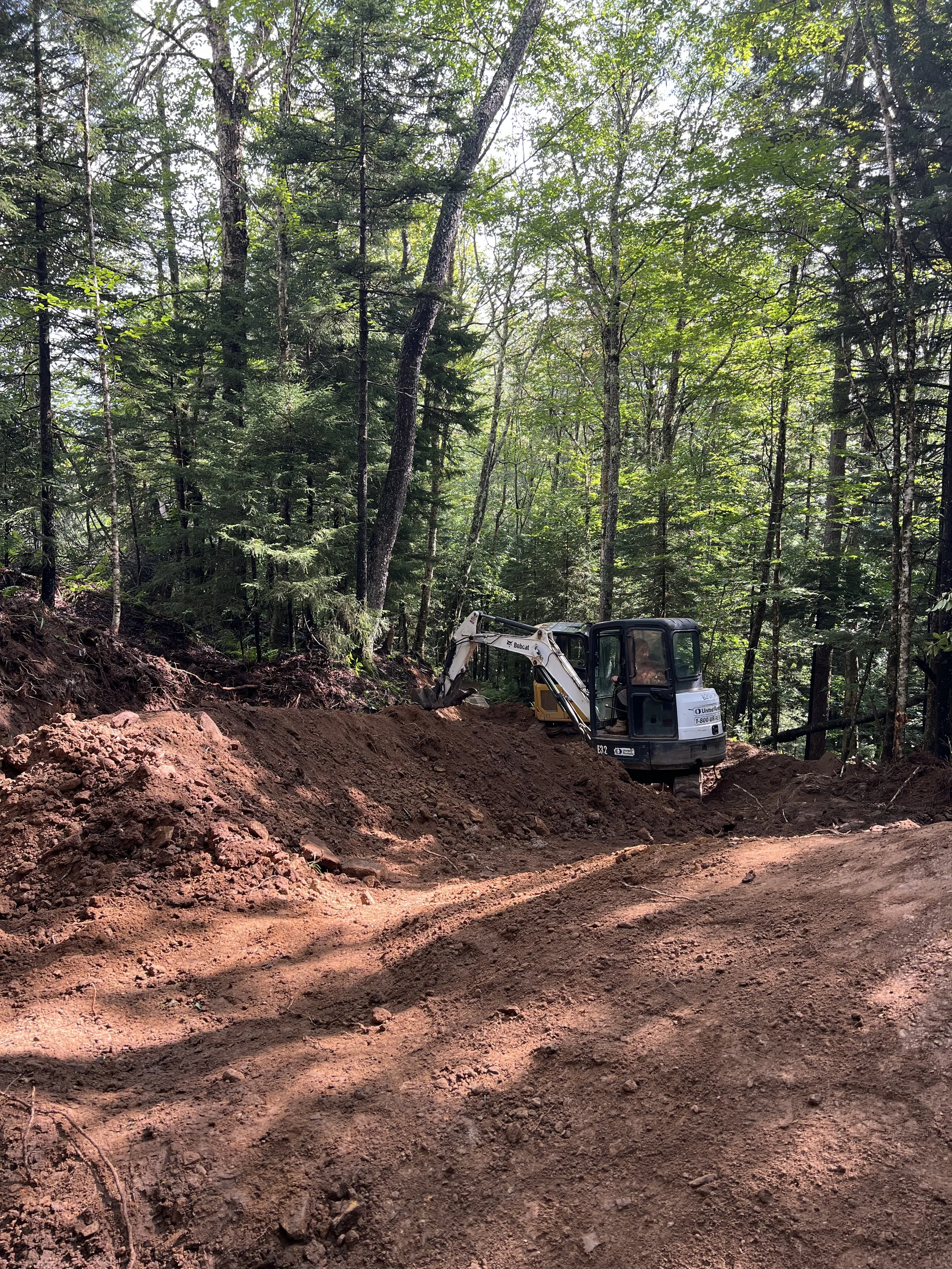 A small excavator working on a dirt trail in a dense forest with tall trees and green leaves.