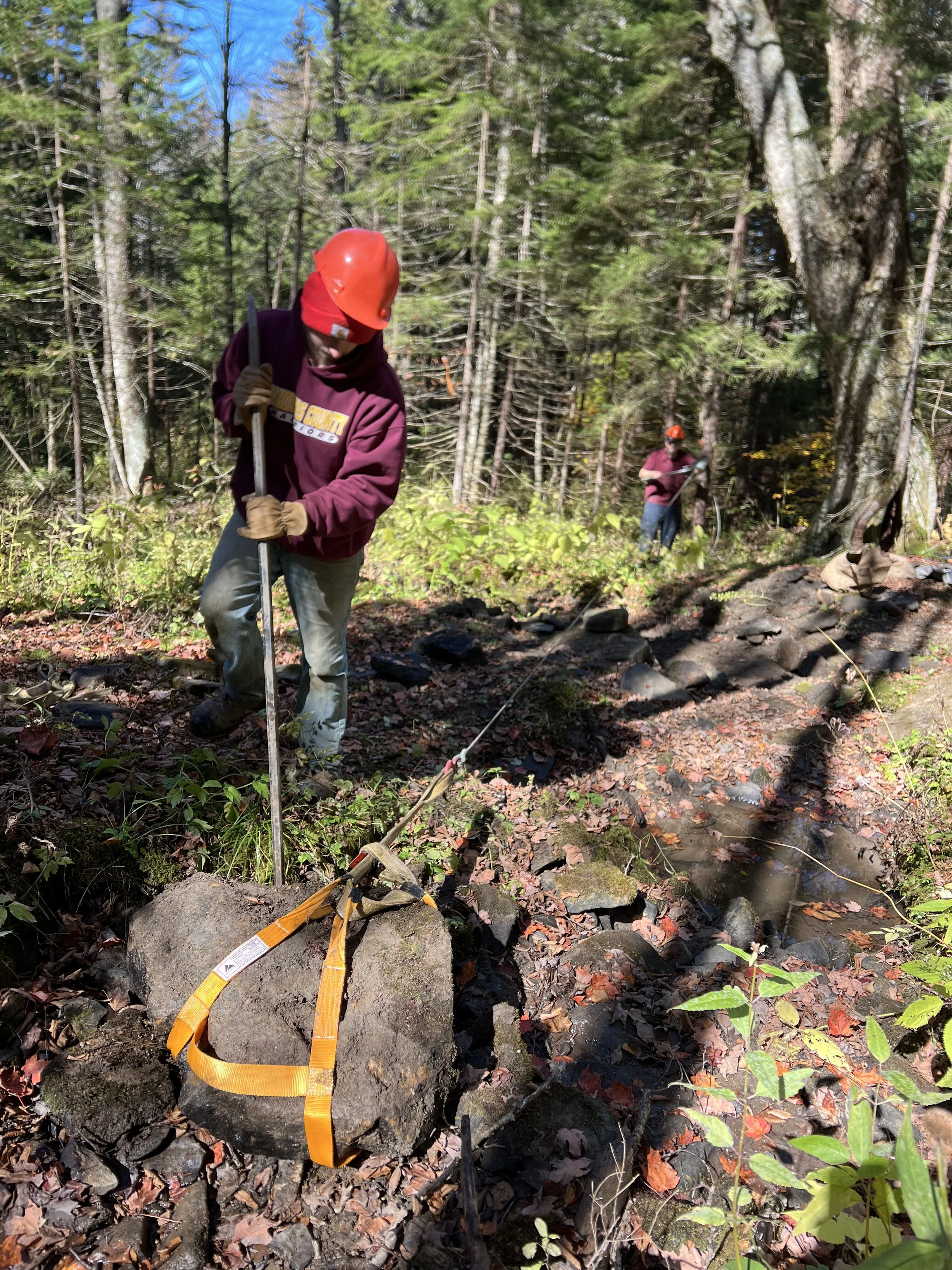 Two people working outdoors in a wooded area, installing a rock anchor with safety straps on a large rock near a small stream, both wearing helmets and gloves.