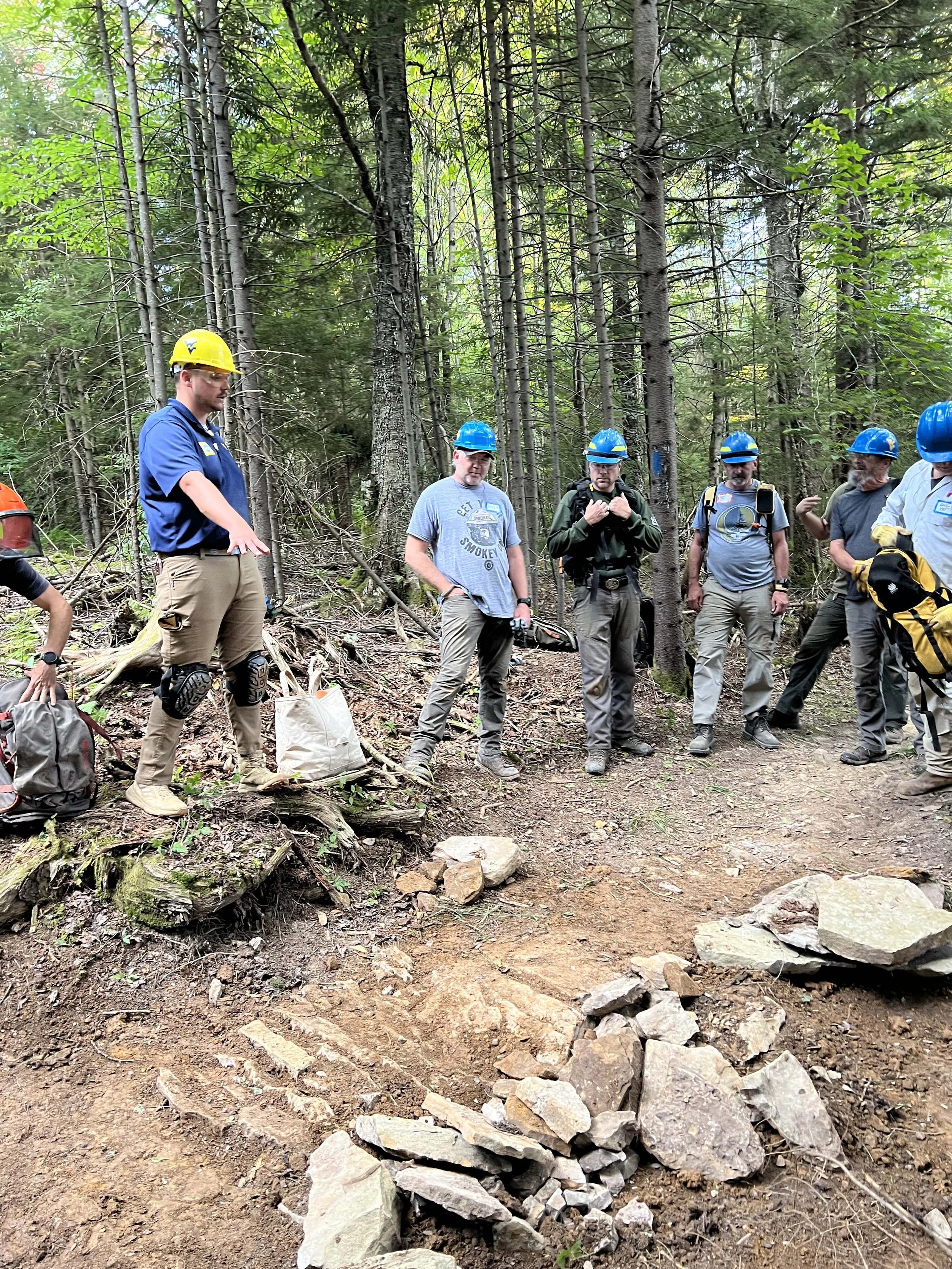 Group of people wearing helmets and safety gear standing in a forested area with a dirt trail and a rock formation in the foreground.