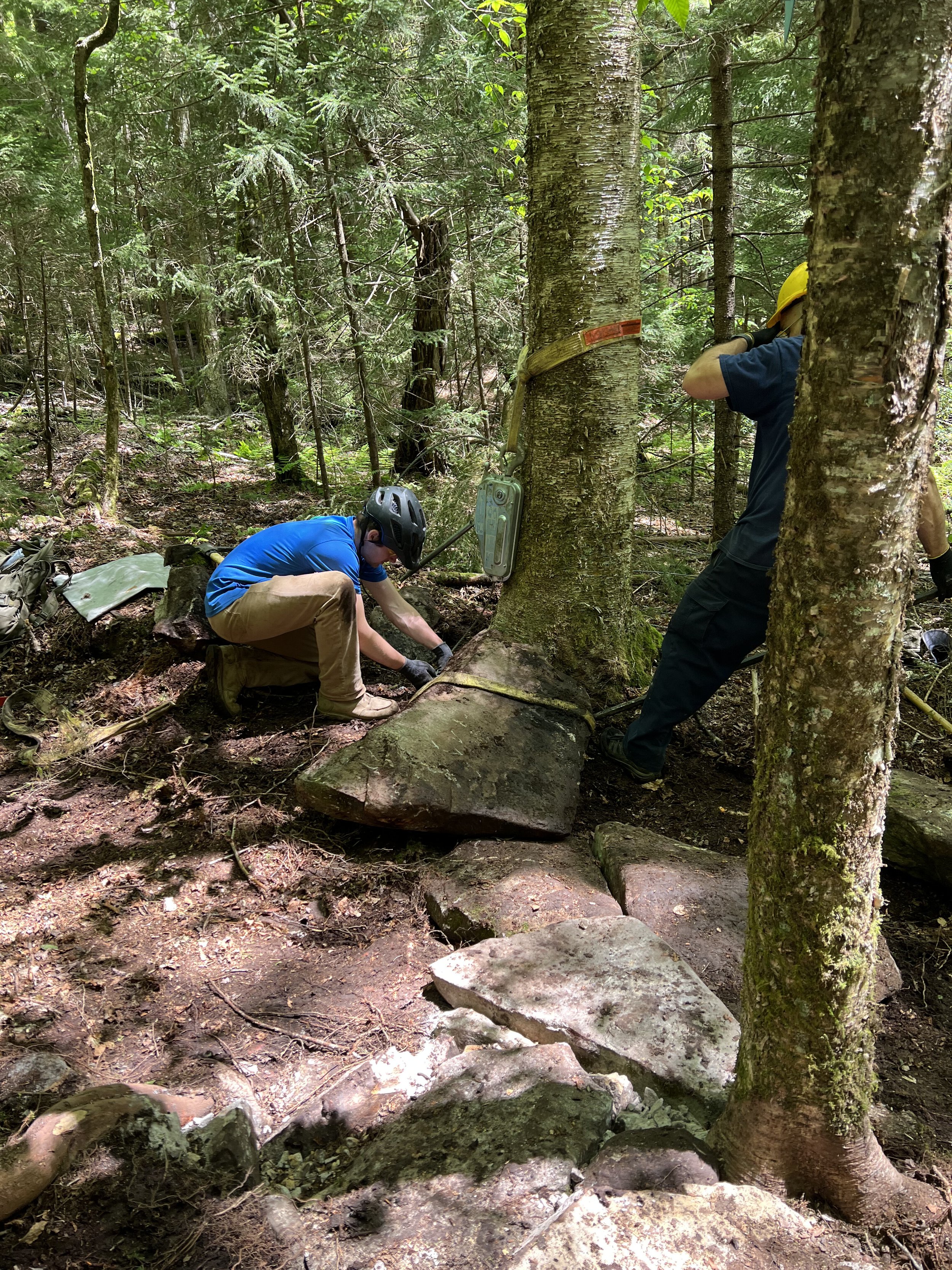 Two workers with helmets working on a large rock in a forest, securing it with straps for transportation or stabilization.