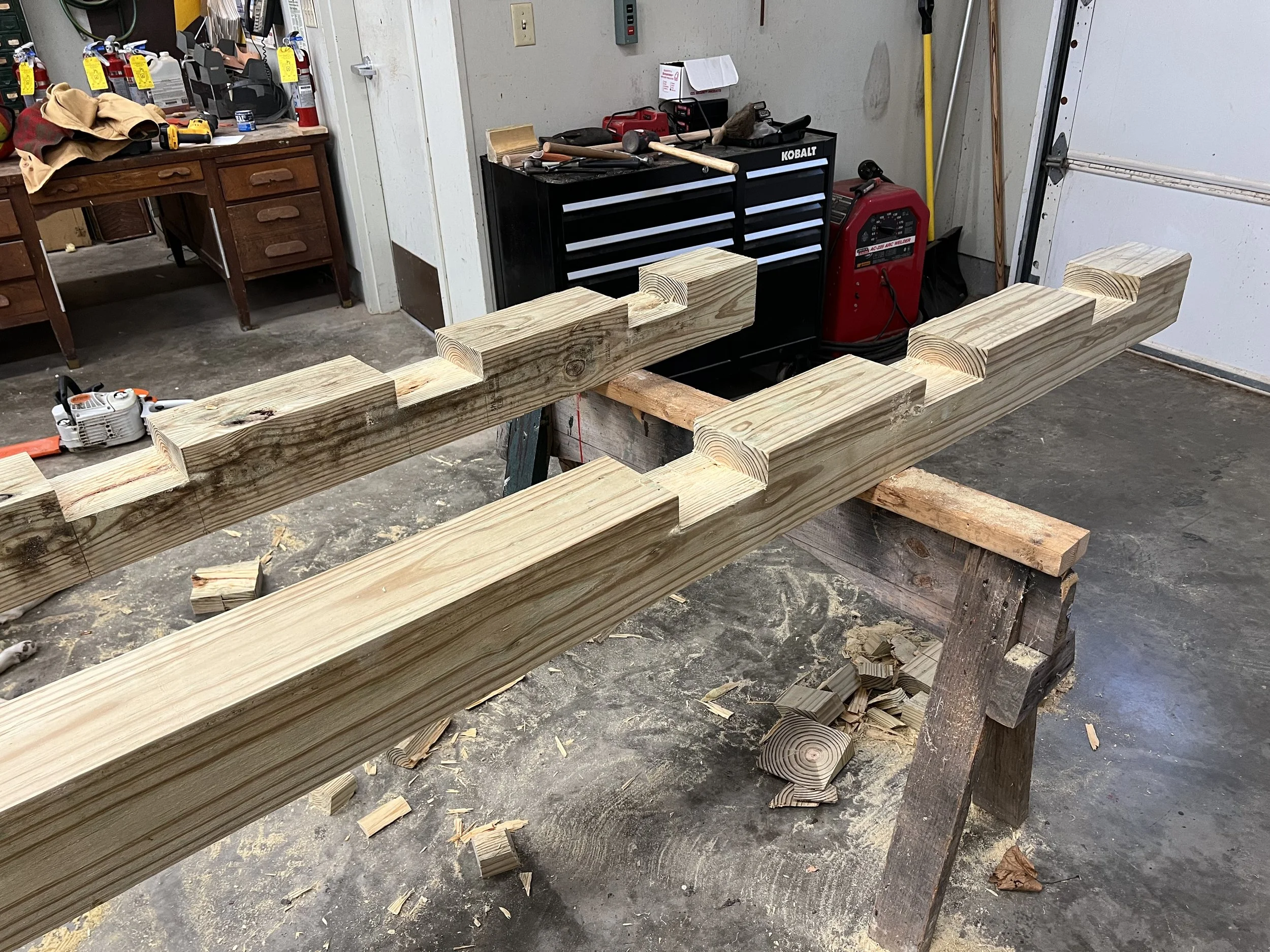Woodworking workshop with pieces of cut lumber on a workbench, a chainsaw on the floor, tools and a tool chest in the background.