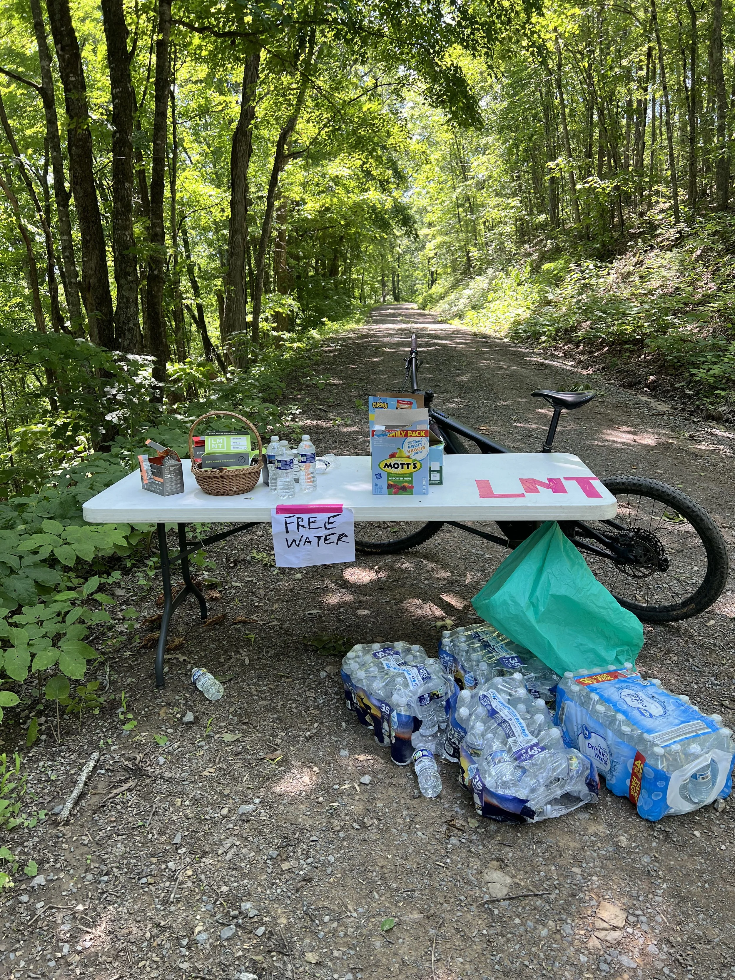 A water station set up along a wooded forest trail with bottled water, snacks, and a sign offering free water.