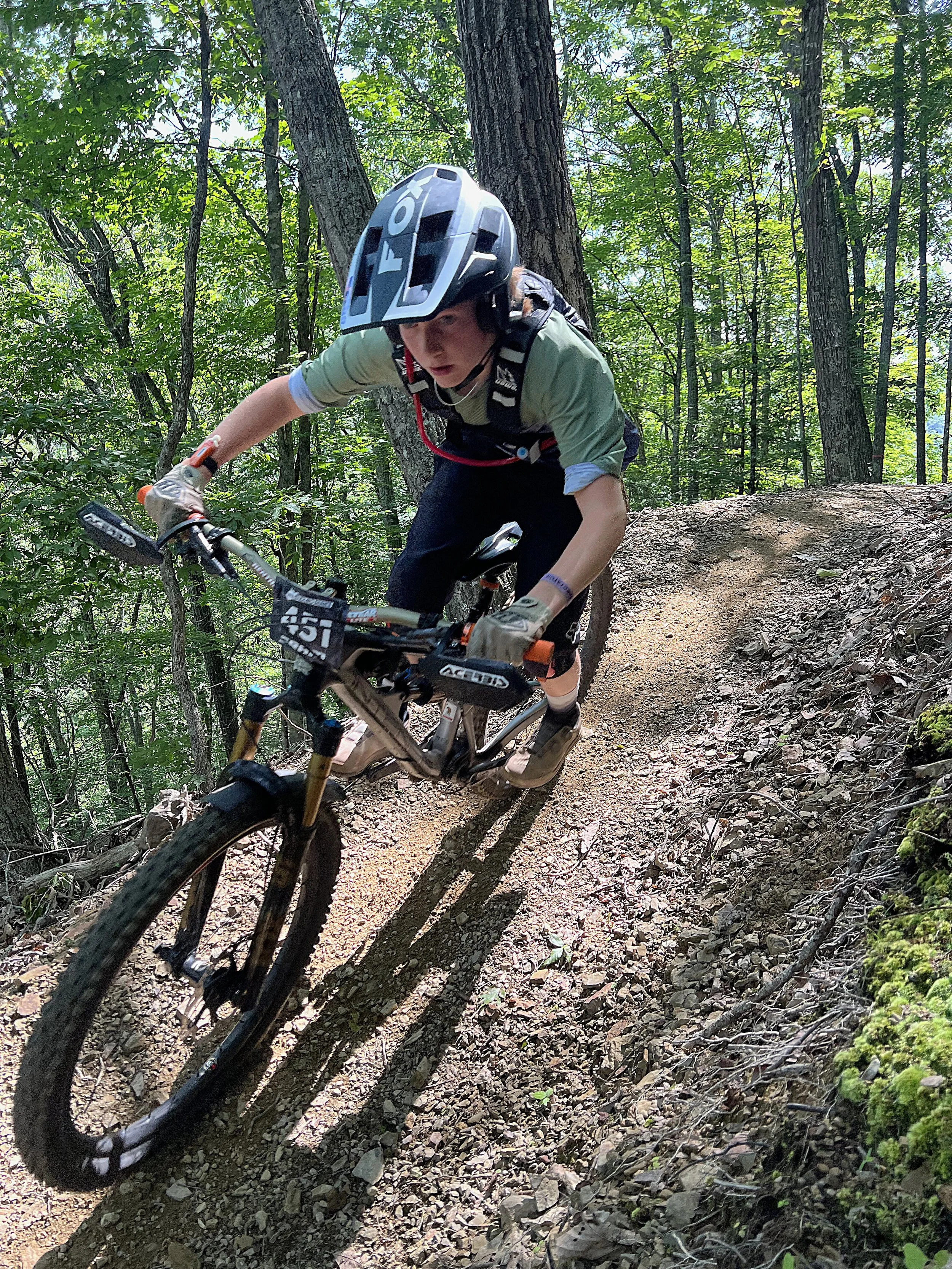 A person mountain biking on a dirt trail through a forest, wearing a helmet, gloves, and protective gear.