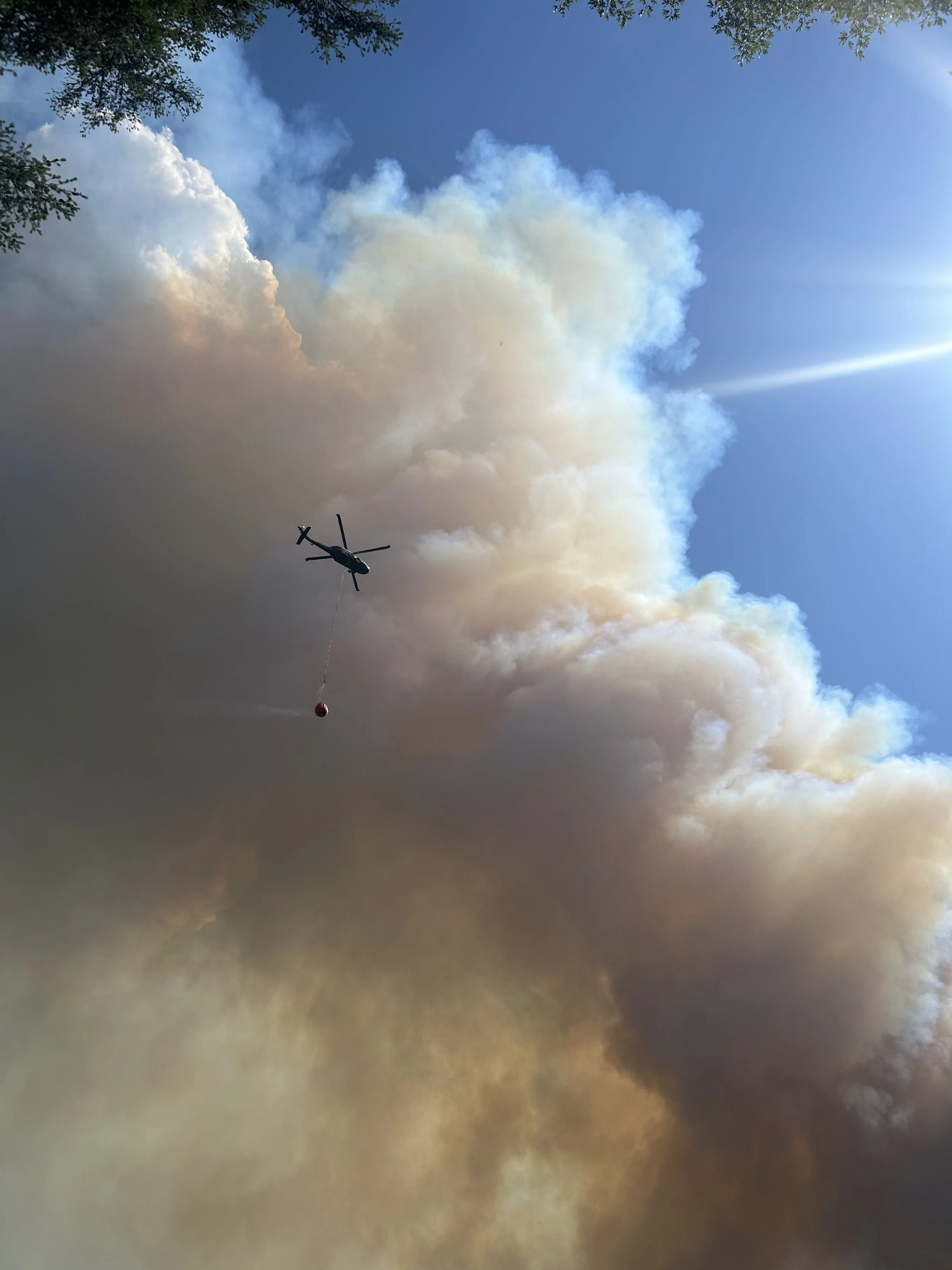 Helicopter dropping water to combat a wildfire with thick smoke and blue sky in the background.