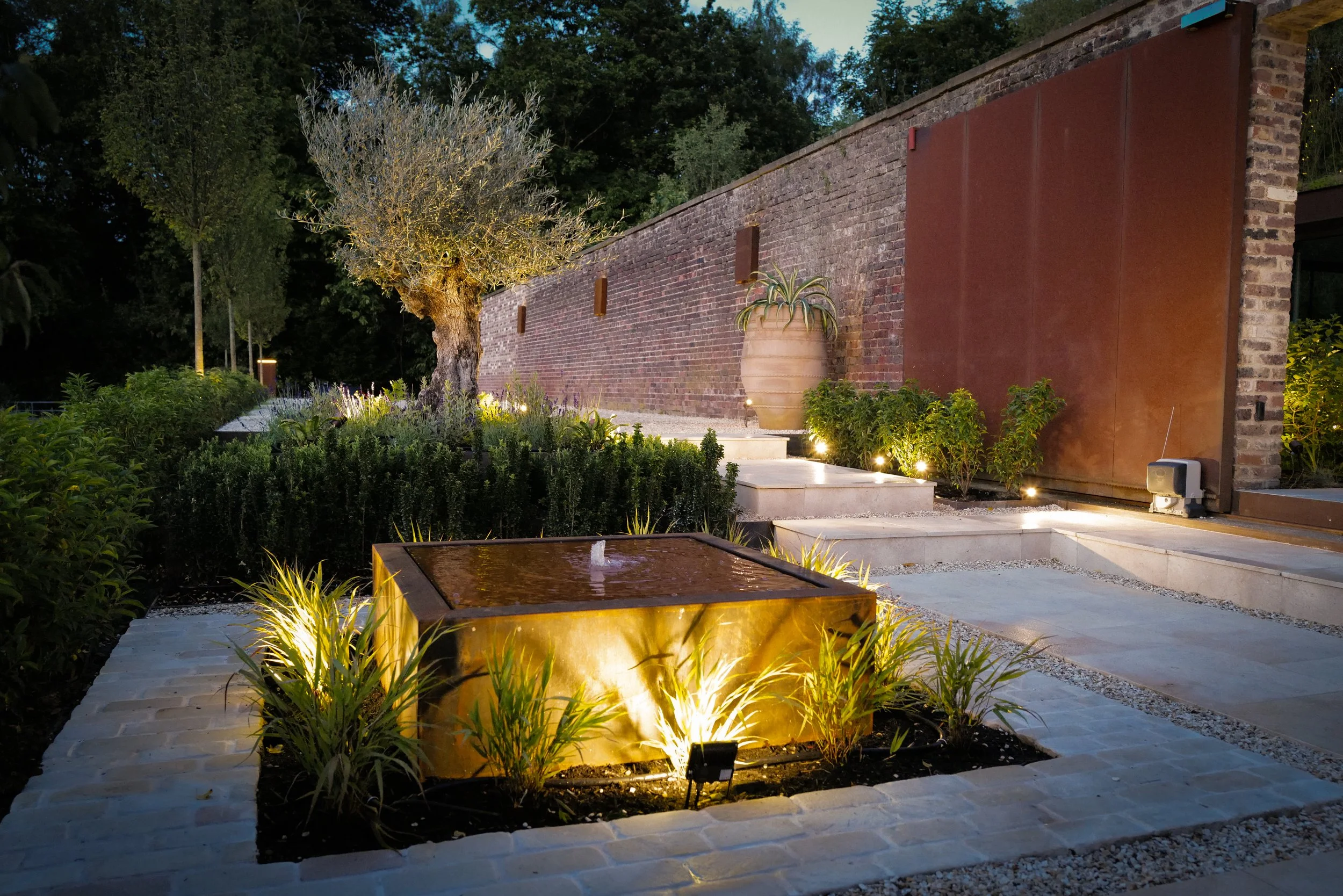 A landscaped outdoor garden area at night with illuminated plants, a small water feature, a brick wall, and outdoor lighting.