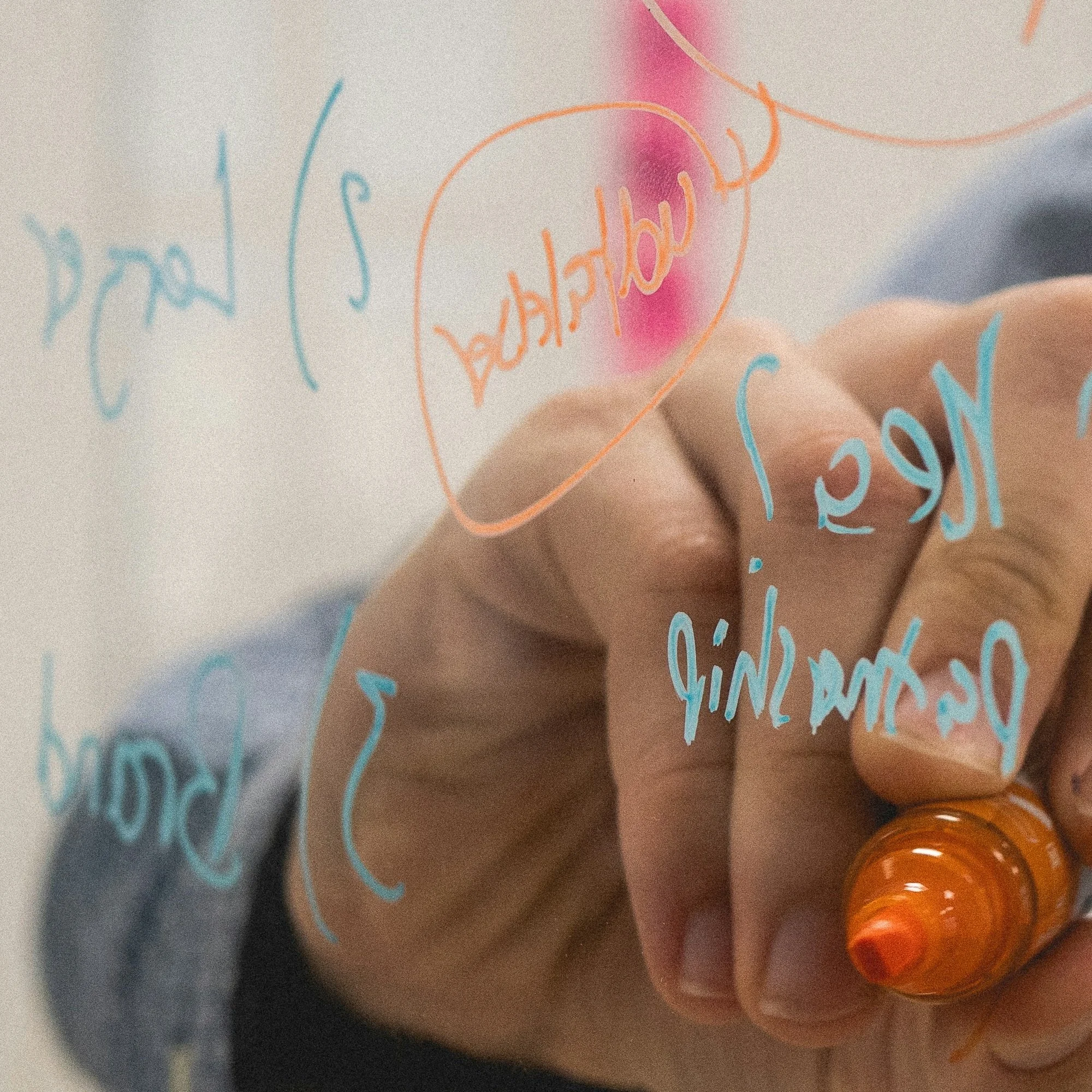 Person writing on a transparent surface with colorful markers, words are visible but reversed, including 'Birthday' in red, among other words in blue and orange.