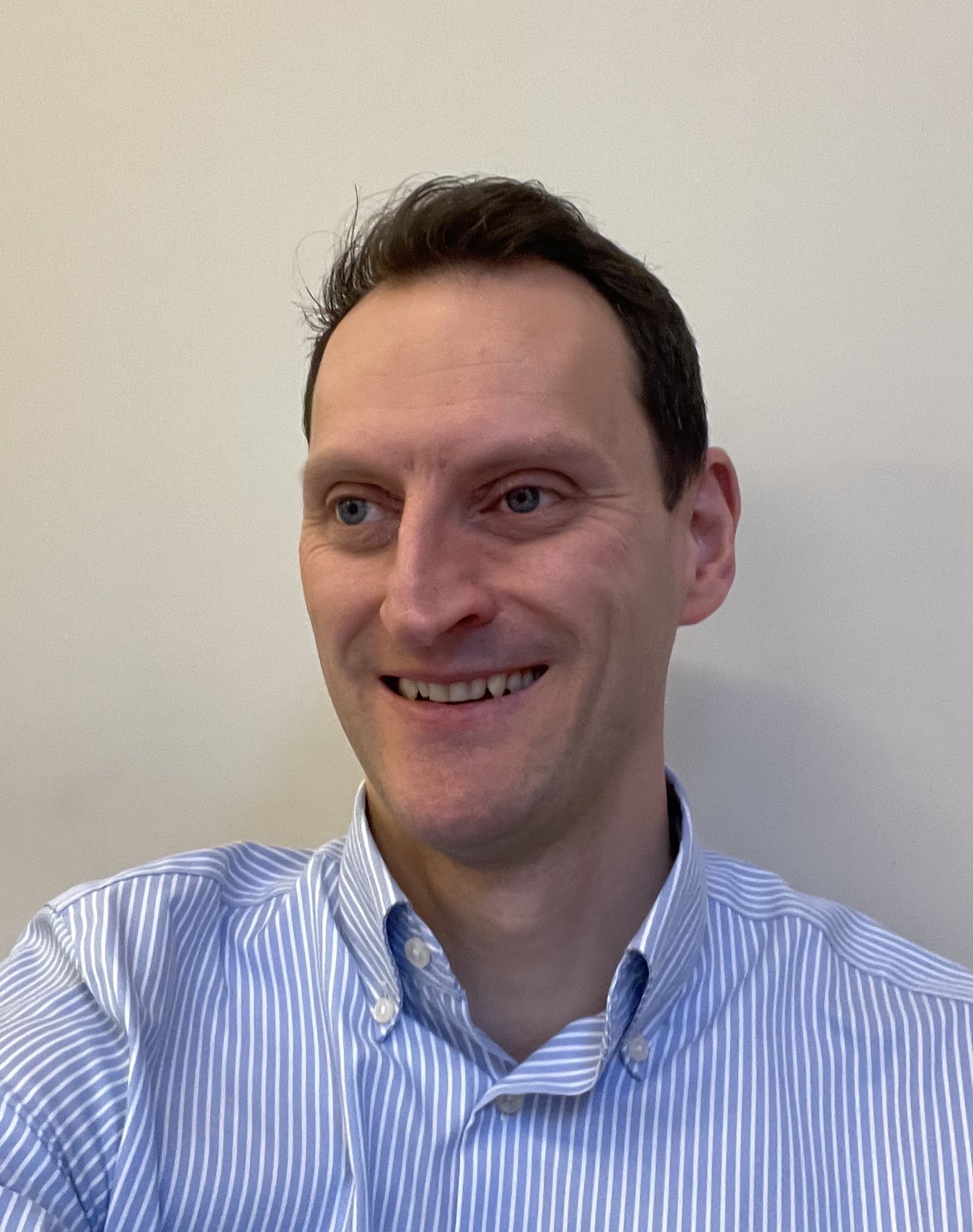A man with short dark hair, wearing a blue and white striped button-up shirt, smiling and looking to the side against a plain light-colored wall.