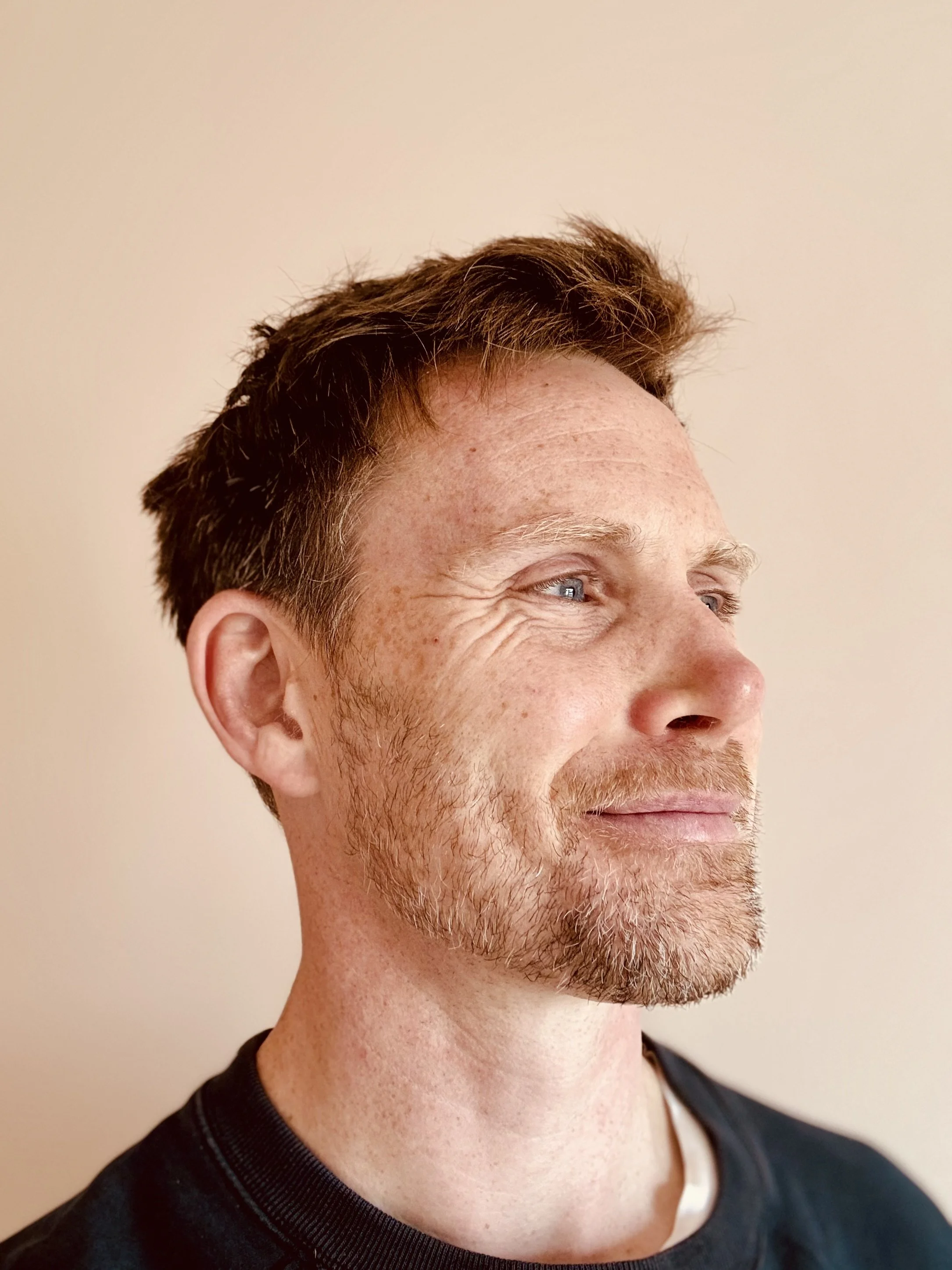 Close-up of a smiling man with light skin, brown hair, blue eyes, and a beard, looking to the side against a plain off-white background.