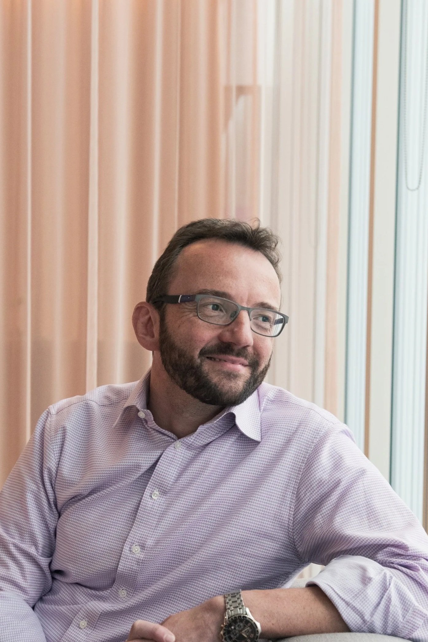 A man with glasses and a beard, smiling and looking to his right, sitting in front of beige curtains and bright window light.