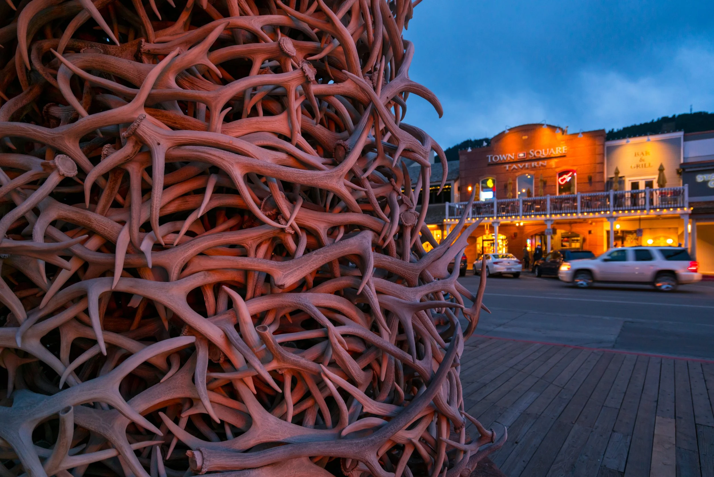 A large sculpture made of numerous intertwined antlers in the foreground with a town square and buildings illuminated at dusk in the background.