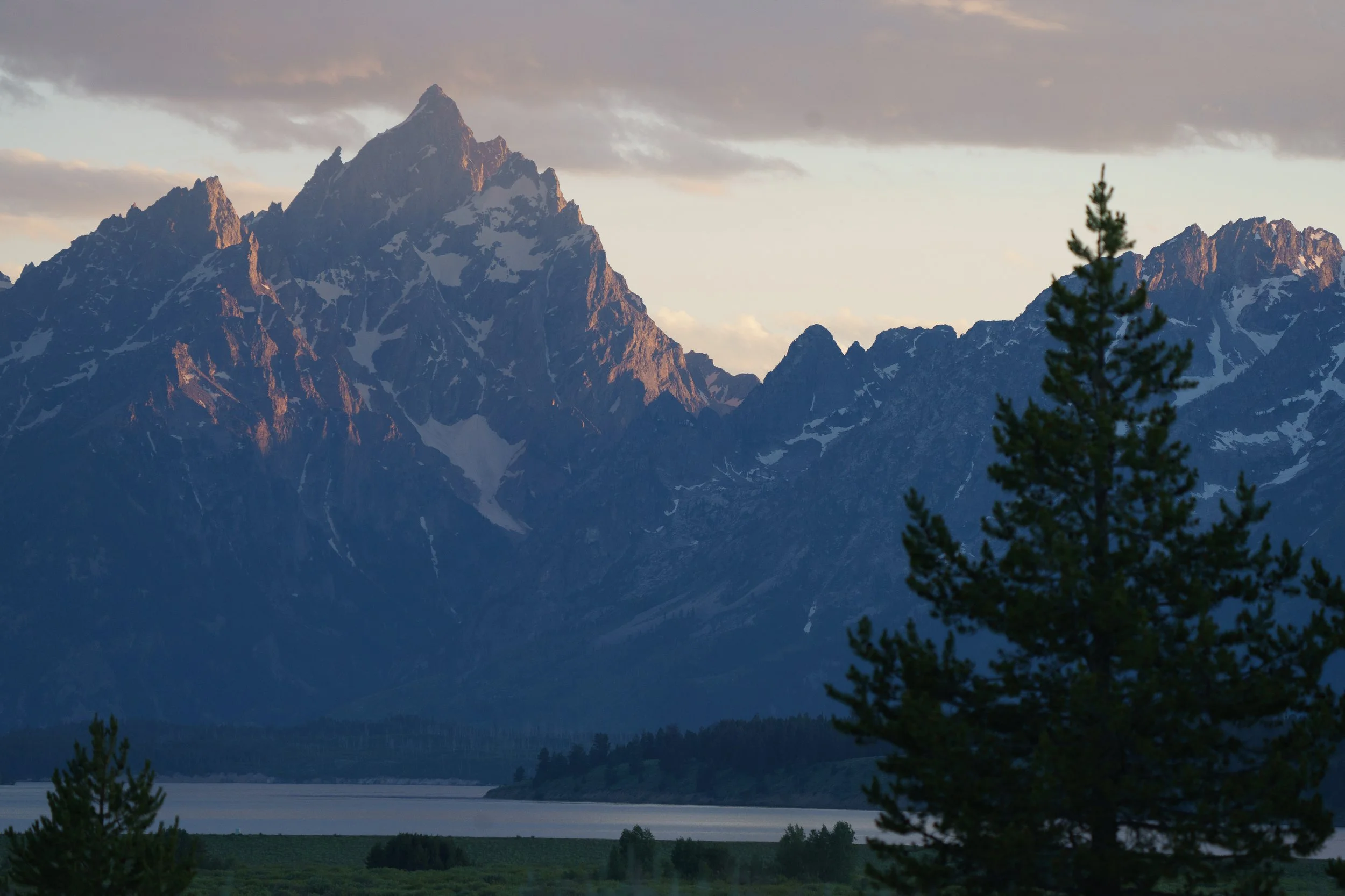 A scenic mountain landscape featuring a large snow-capped mountain with jagged peaks, surrounded by smaller mountains, with a lake and lush green trees in the foreground.
