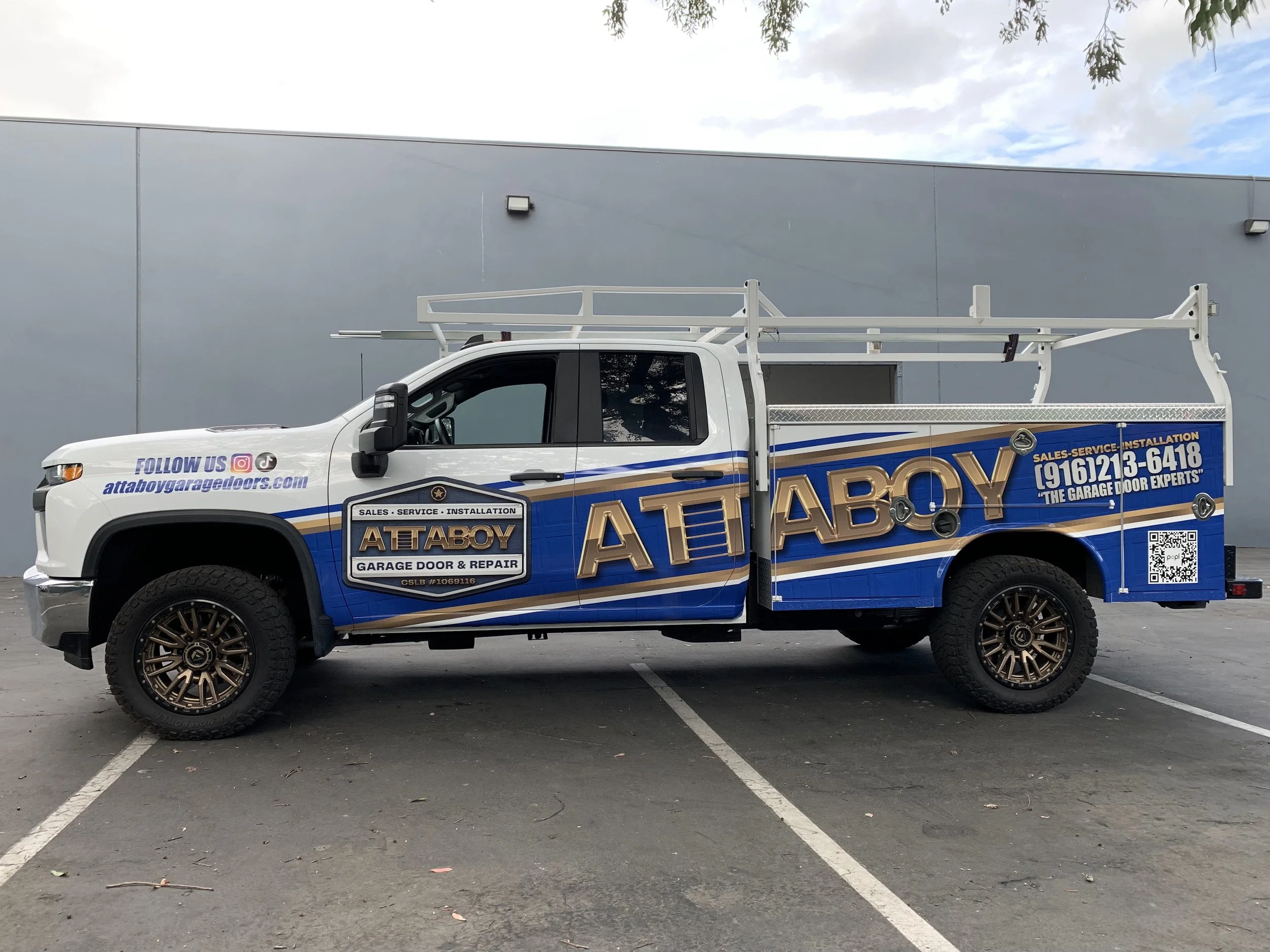 White service truck with blue and gold branding for ATTABOY Garage Door & Repair, parked in an outdoor lot with a gray wall and cloudy sky in the background, featuring a ladder rack on top and contact information and QR code on the side.