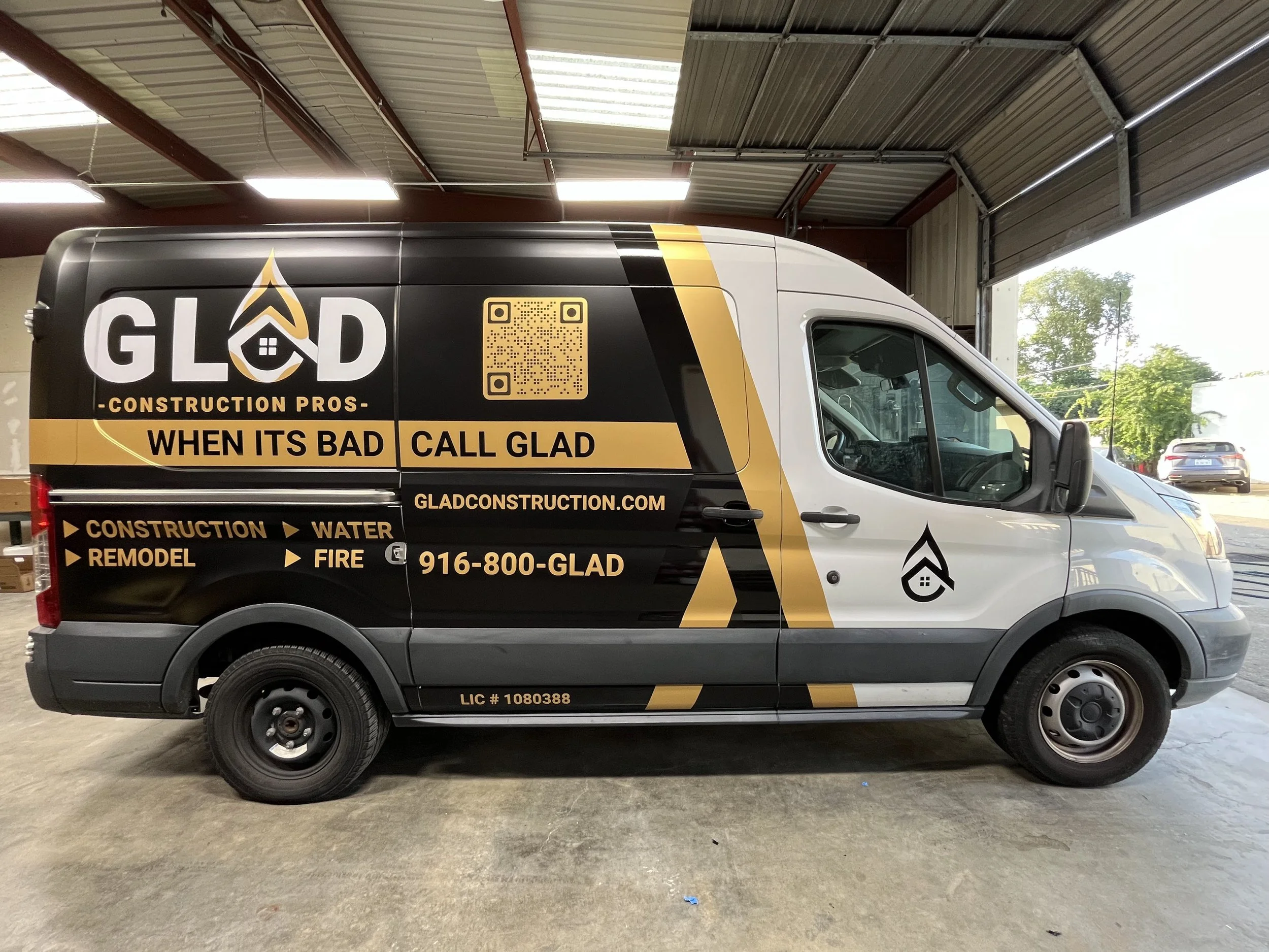 A construction company's van parked inside a warehouse with advertising for Glad Construction Pros, featuring black and gold colors, a QR code, and the company's contact information.