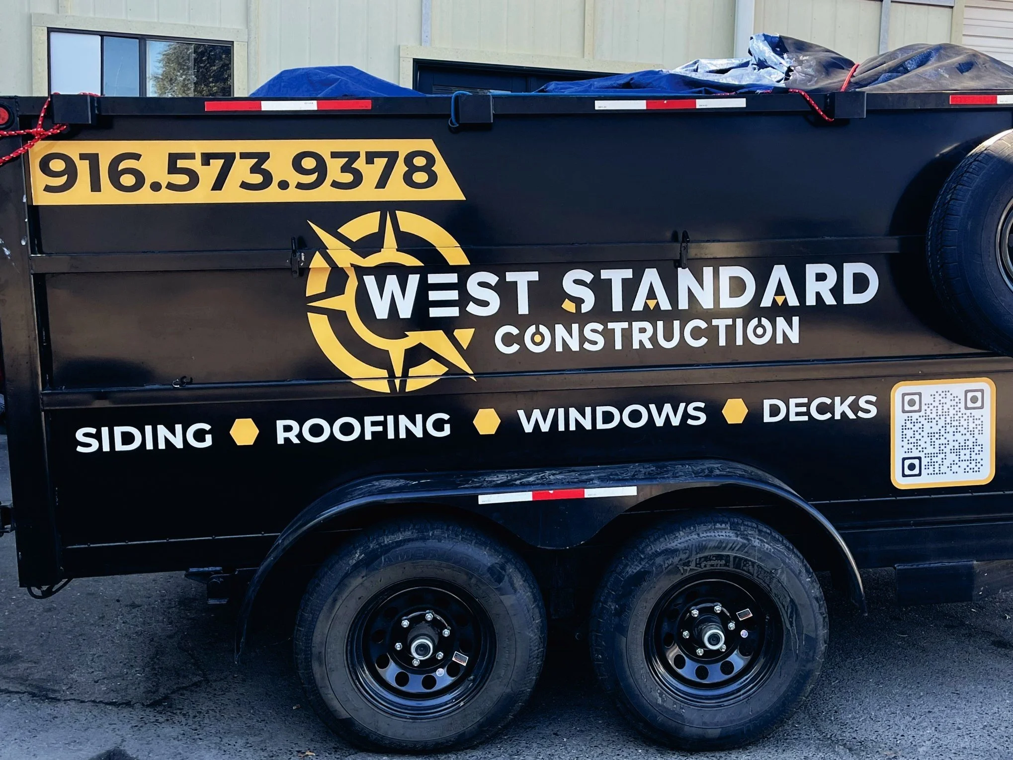 A black construction trailer with yellow and white lettering advertising West Standard Construction. The trailer features a phone number 916.573.9378, a logo resembling a compass with a star, and lists services including siding, roofing, windows, and decks. There is a QR code on the bottom right corner of the trailer.