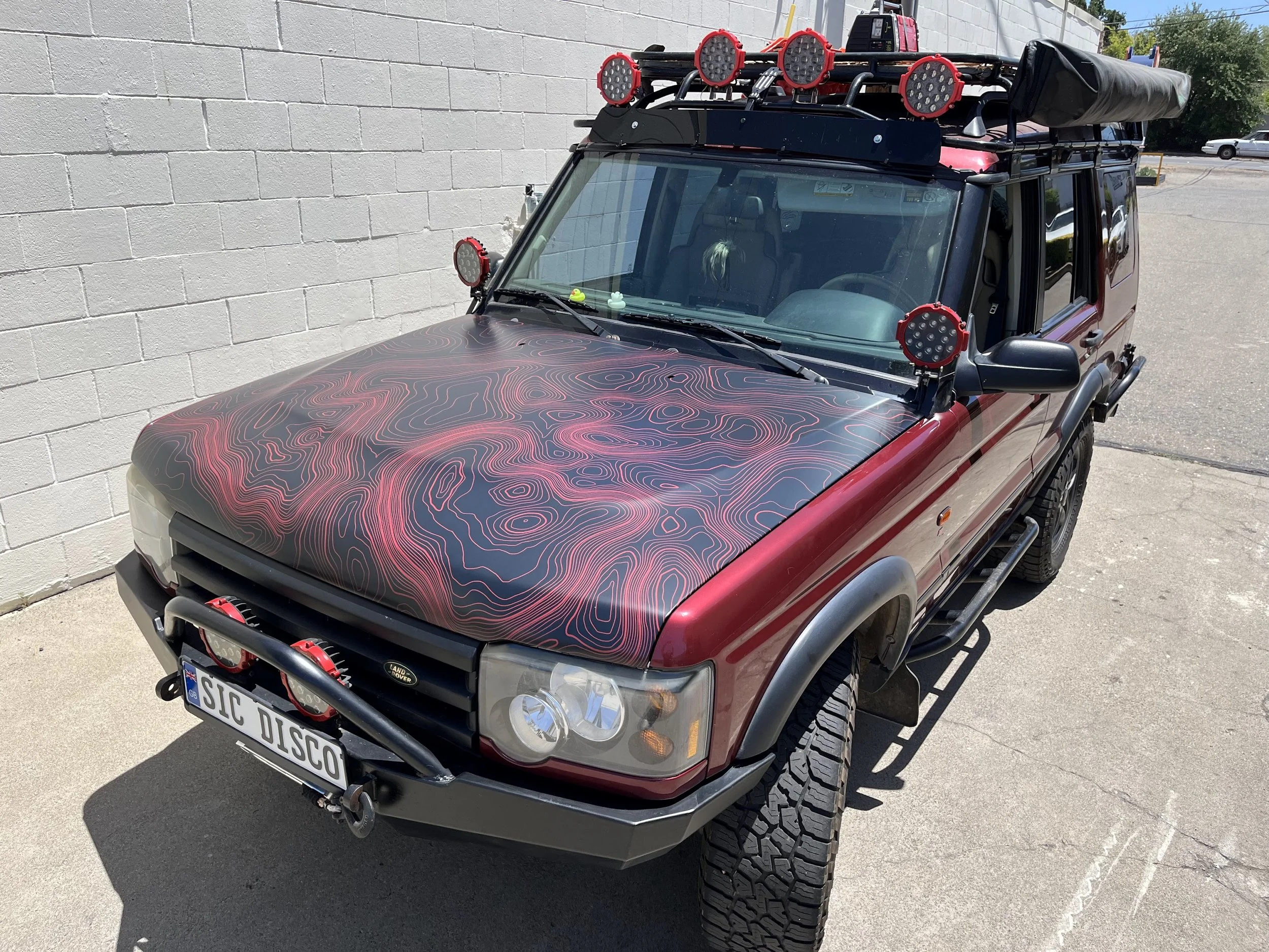 A red Land Rover Defender parked outdoors near a white brick wall, with a custom hood and multiple auxiliary lights mounted on the roof and front bumper.
