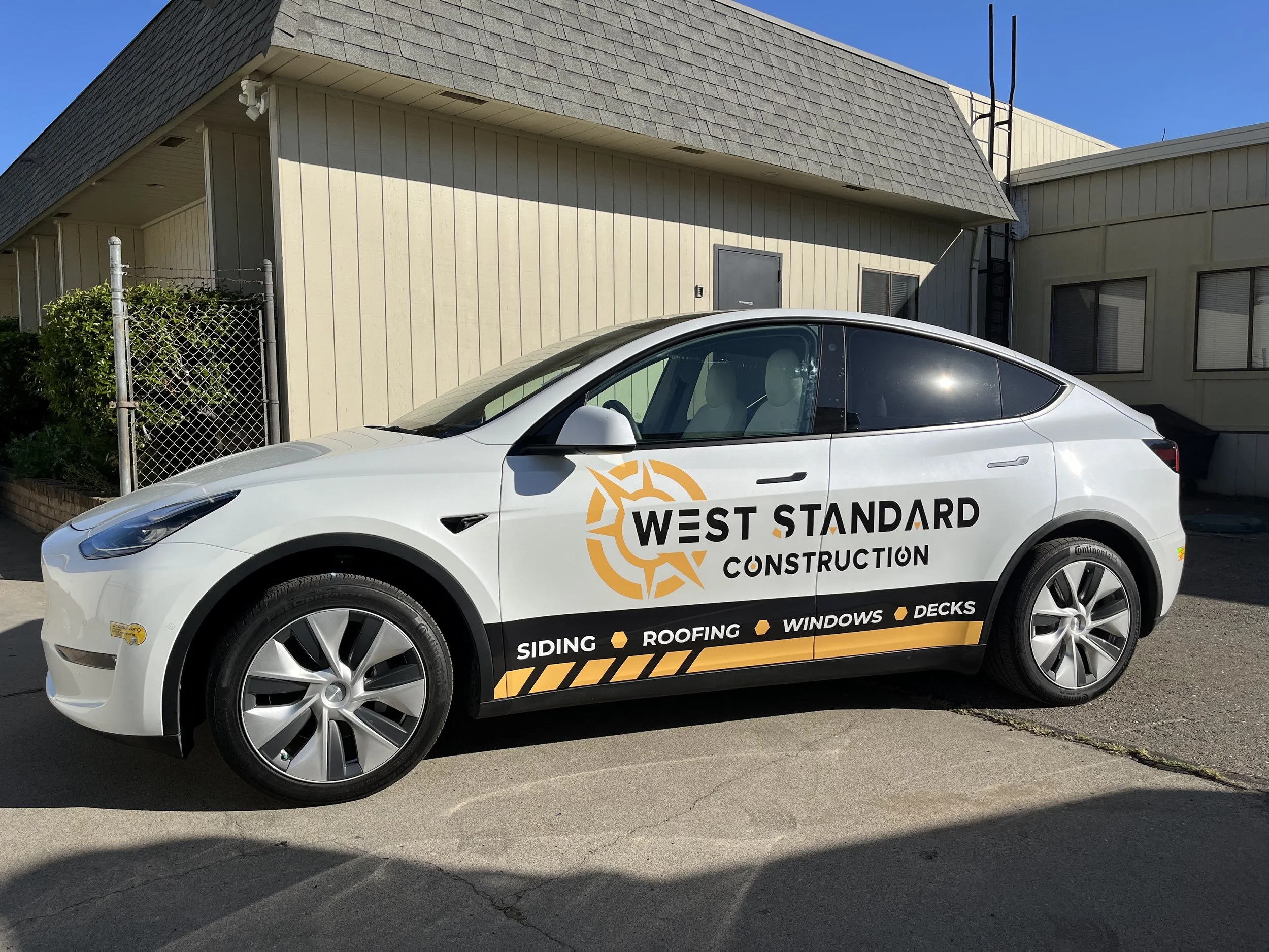 White electric vehicle with West Standard Construction branding parked outdoors in front of a beige building with windows and a chain-link fence.