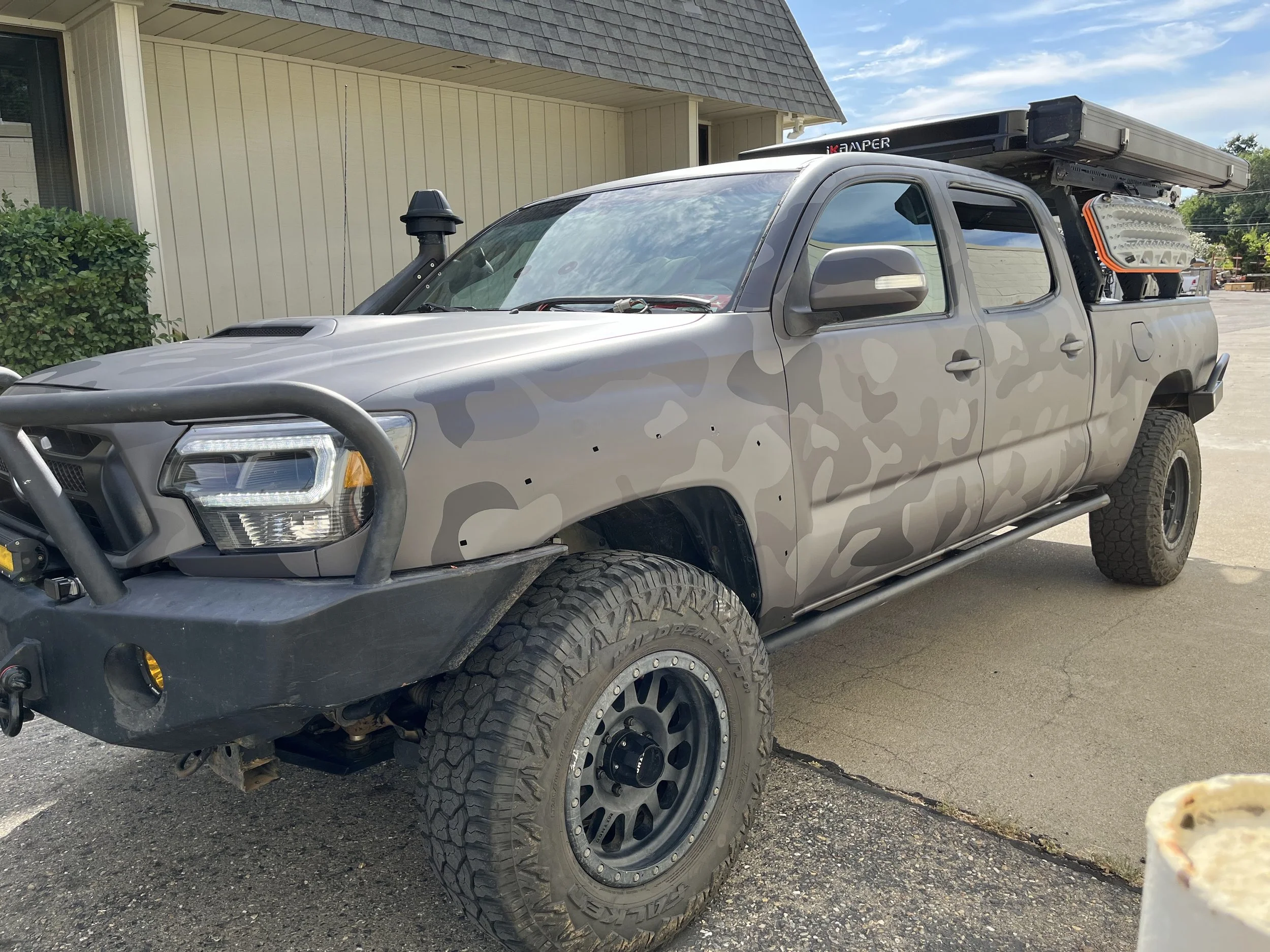A pickup truck with a camouflage paint job, off-road tires, front bumper guard, and utility equipment in the truck bed, parked outside a building with a beige exterior.