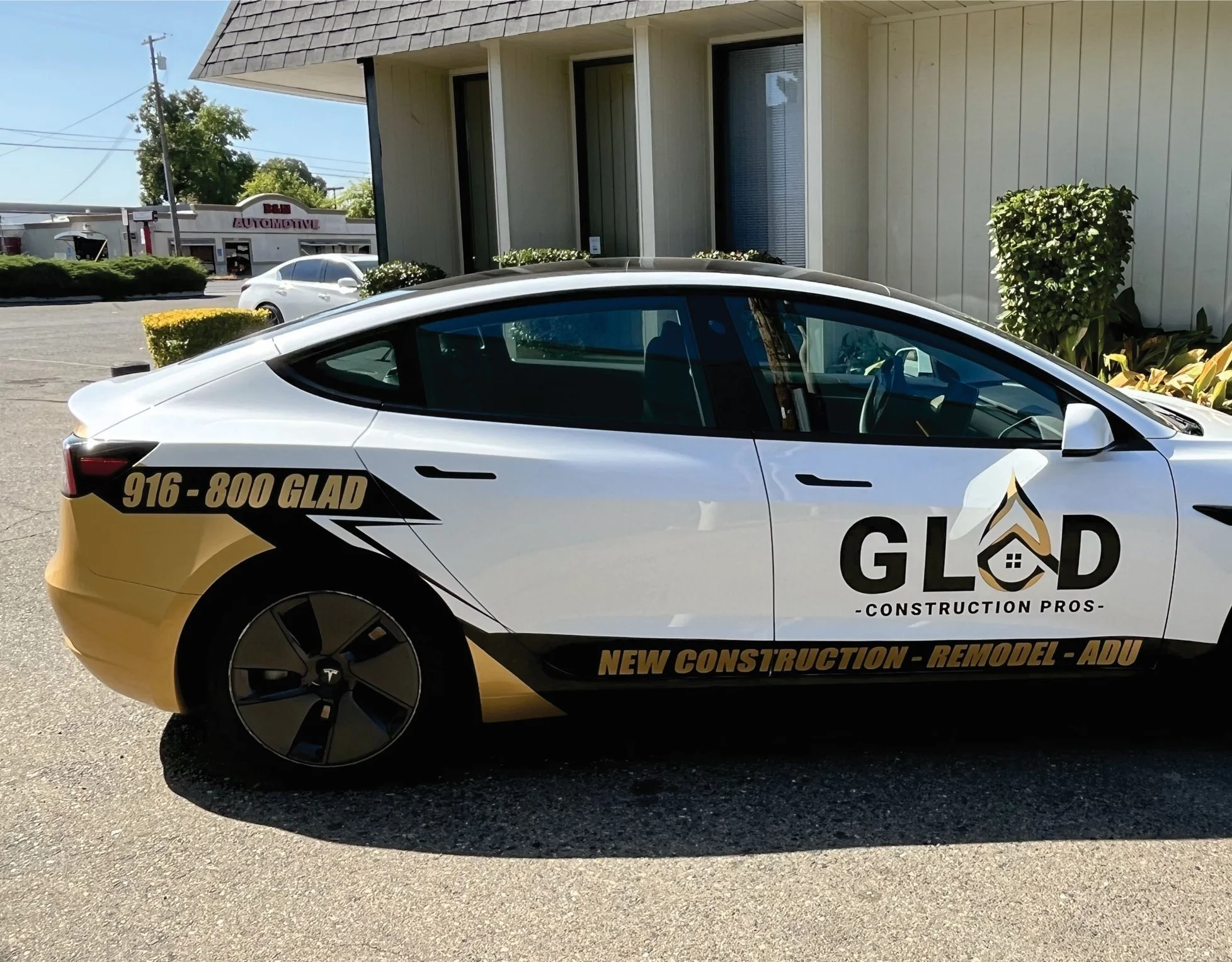 A white Tesla car with black and gold decal graphics for GLOD Construction Pros, featuring a house logo in the shape of a house within the letter G, and the text 'NEW CONSTRUCTION - REMODEL - ADU'. The car is parked outside a building with bushes and another vehicle in the background.
