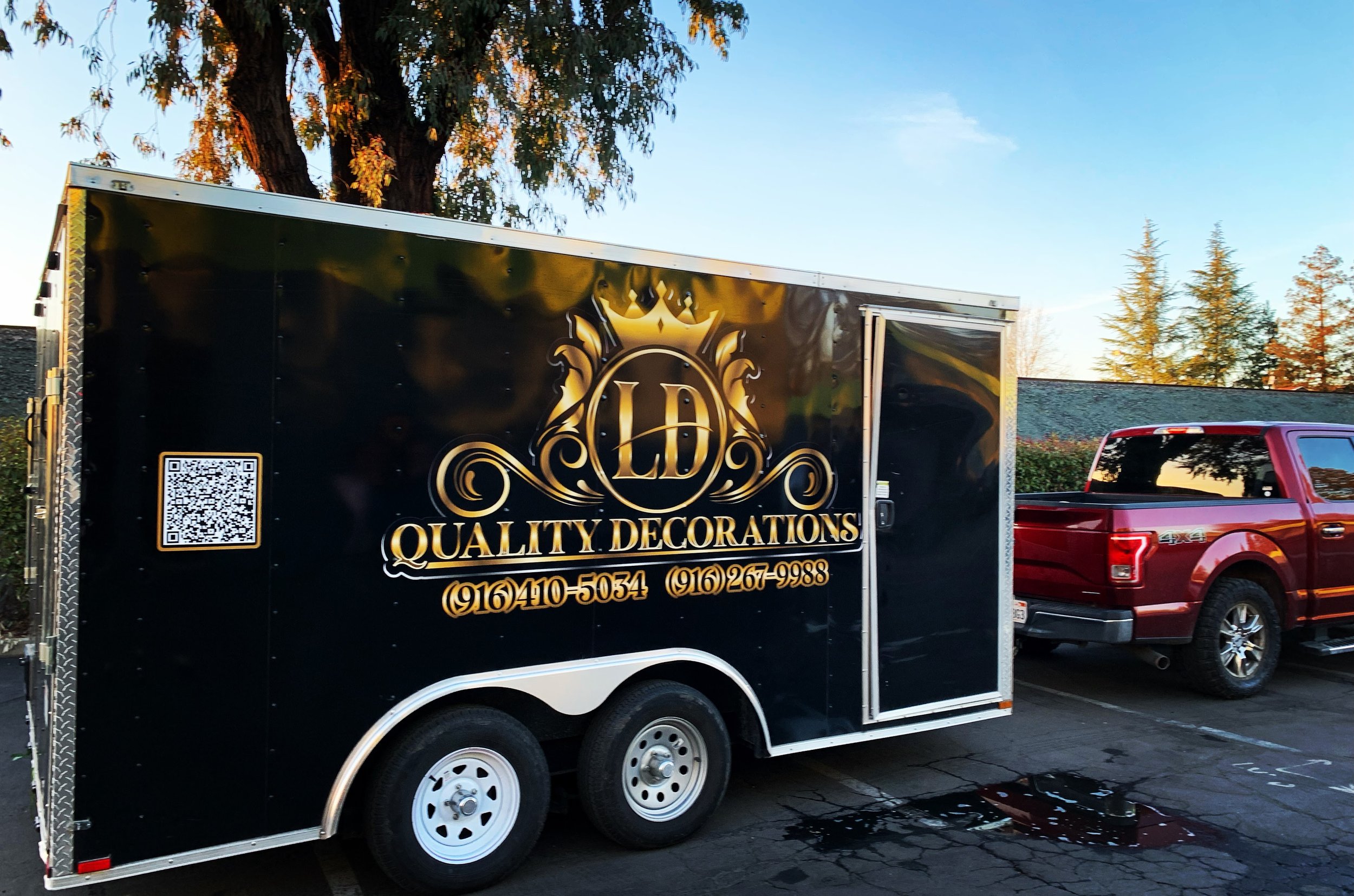 Black trailer with gold lettering and logo labeled 'Quality Decorations' parked in a lot, with a red pickup truck nearby and trees in the background.