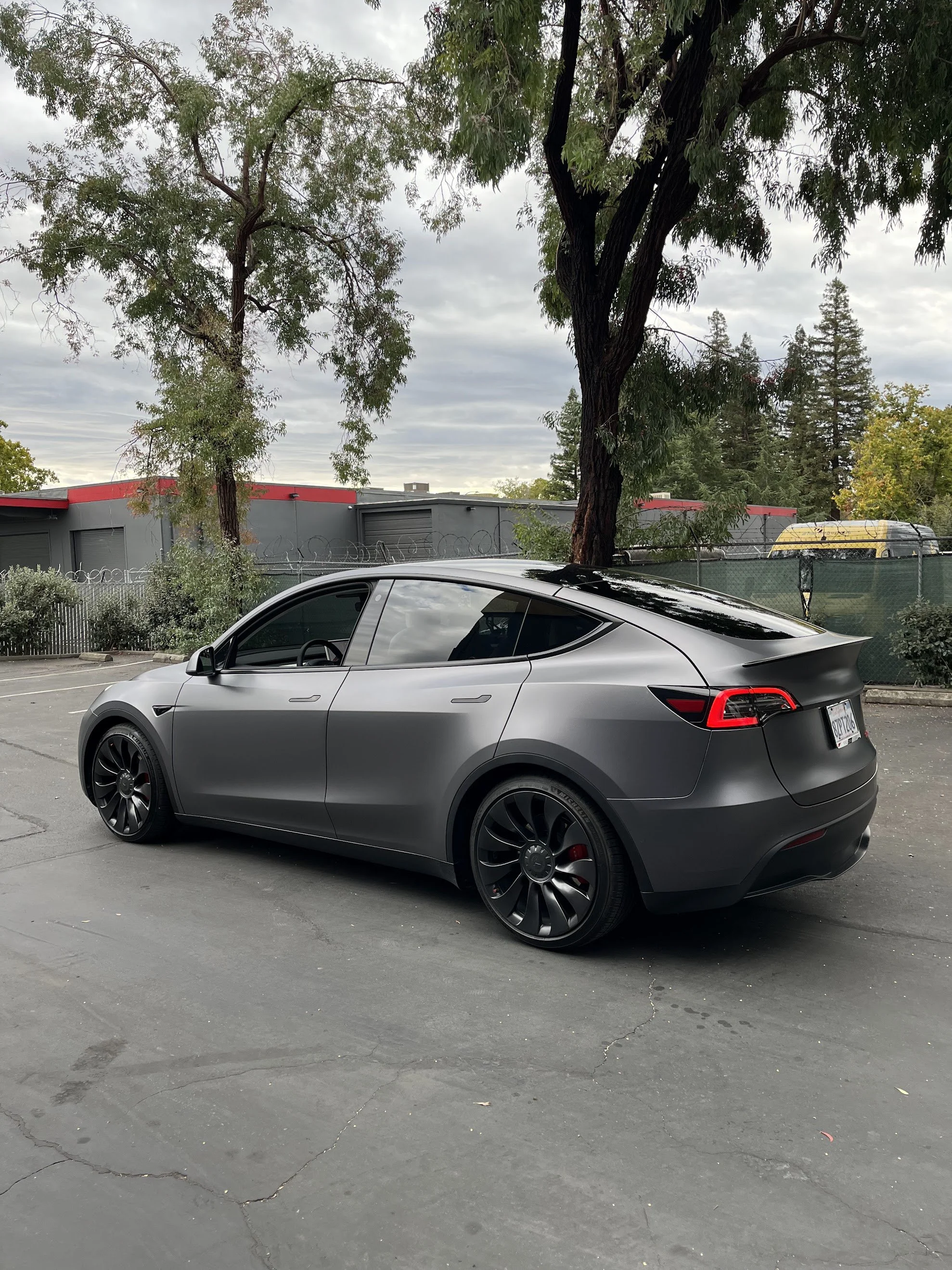 A matte gray Tesla Model Y parked on a concrete surface under trees with green foliage, overcast sky, and a fence in the background.