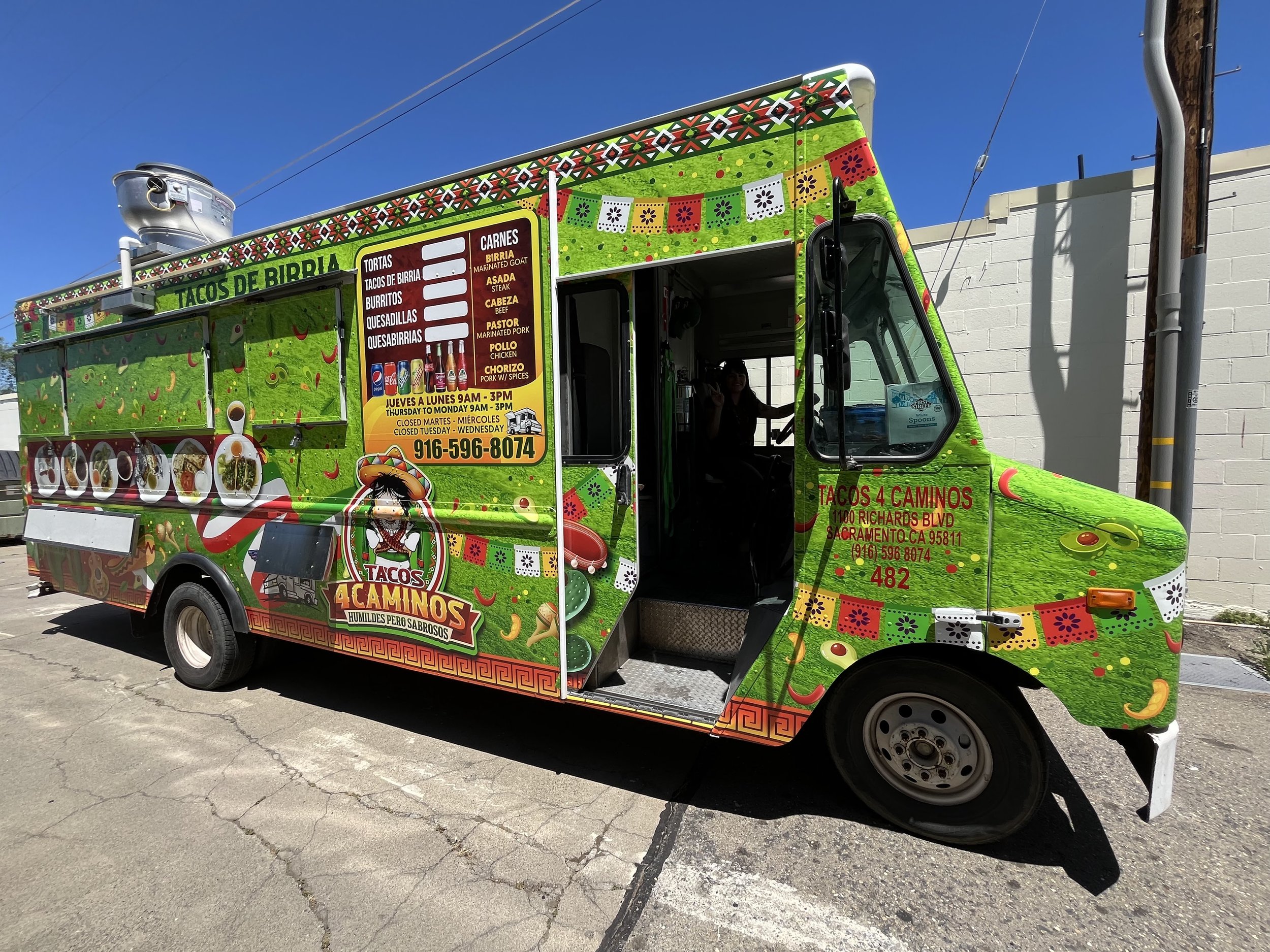 Colorful Mexican taco food truck parked on the street against a blue sky, decorated with vibrant green, red, yellow, and white patterns, advertising tacos, tortas, burritos, quesadillas, and quesabirria.
