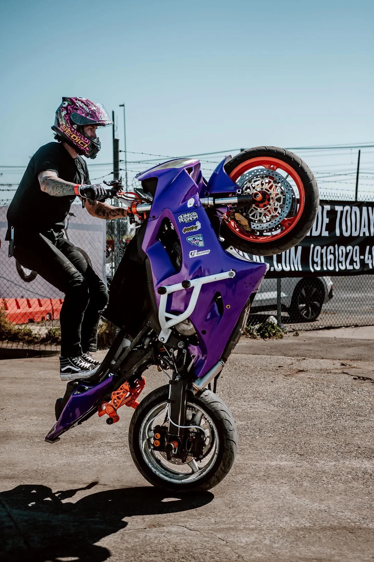 A person wearing a motorcycle helmet and black clothes performing a wheelie stunt on a purple motorcycle in an outdoor area.