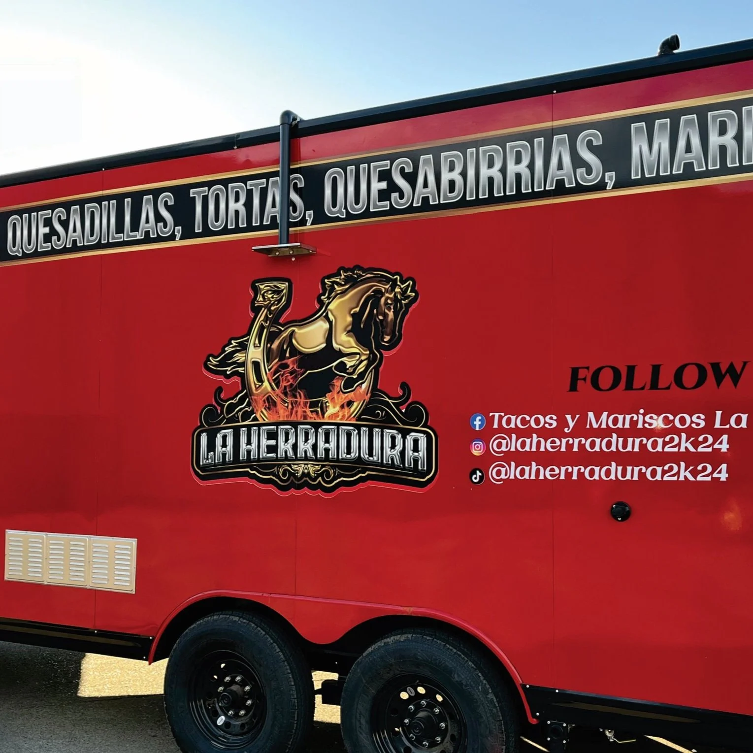 Red food truck with a logo of a galloping horse and flames, advertising Mexican dishes including quesadillas, tacos, and quesabirrias, with social media handles and the name La Herradura.