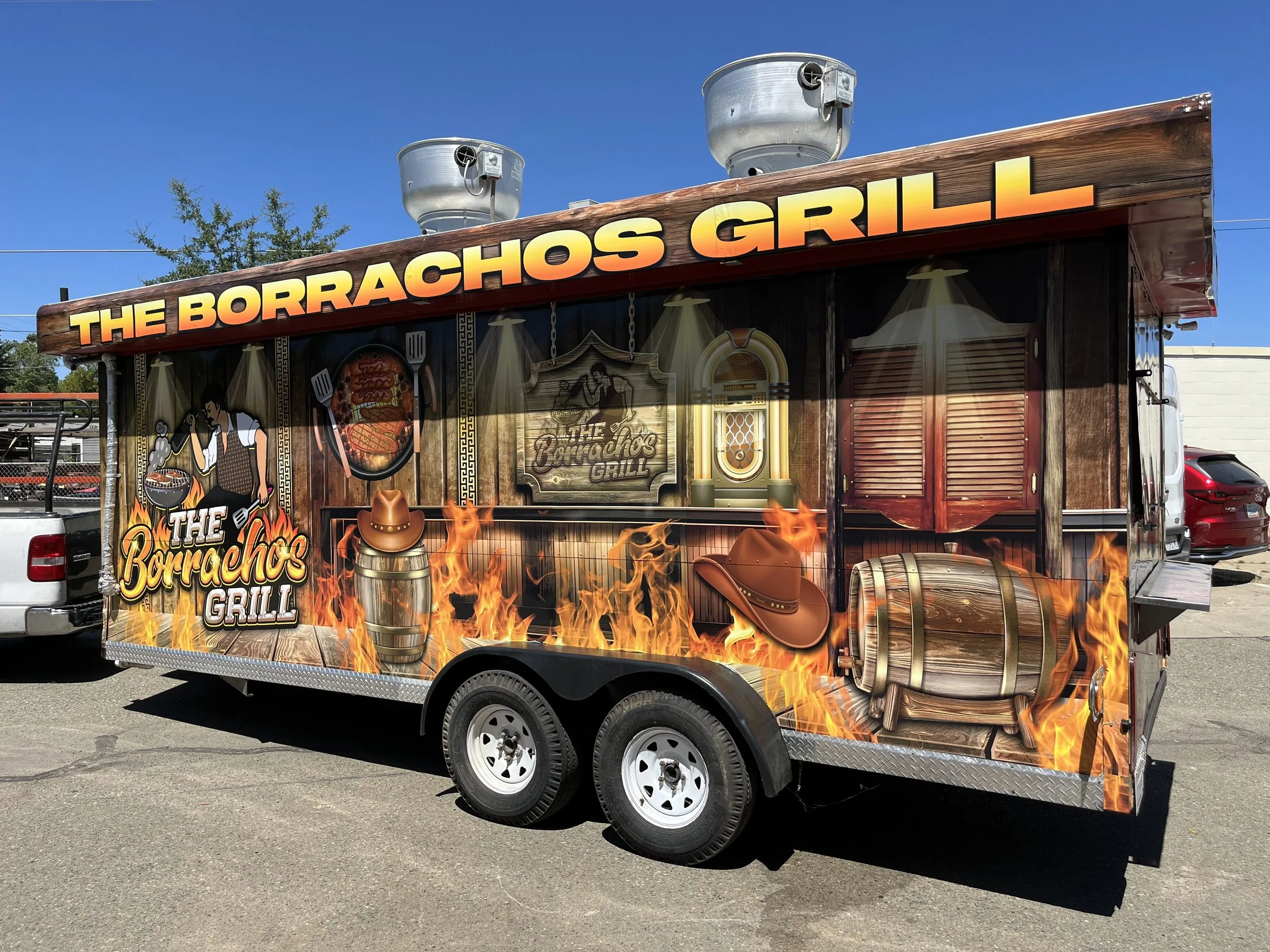 A food trailer with a Wild West theme named 'The Borrachos Grill' featuring flames, cowboy hats, and a barrel, parked outdoors on a sunny day.