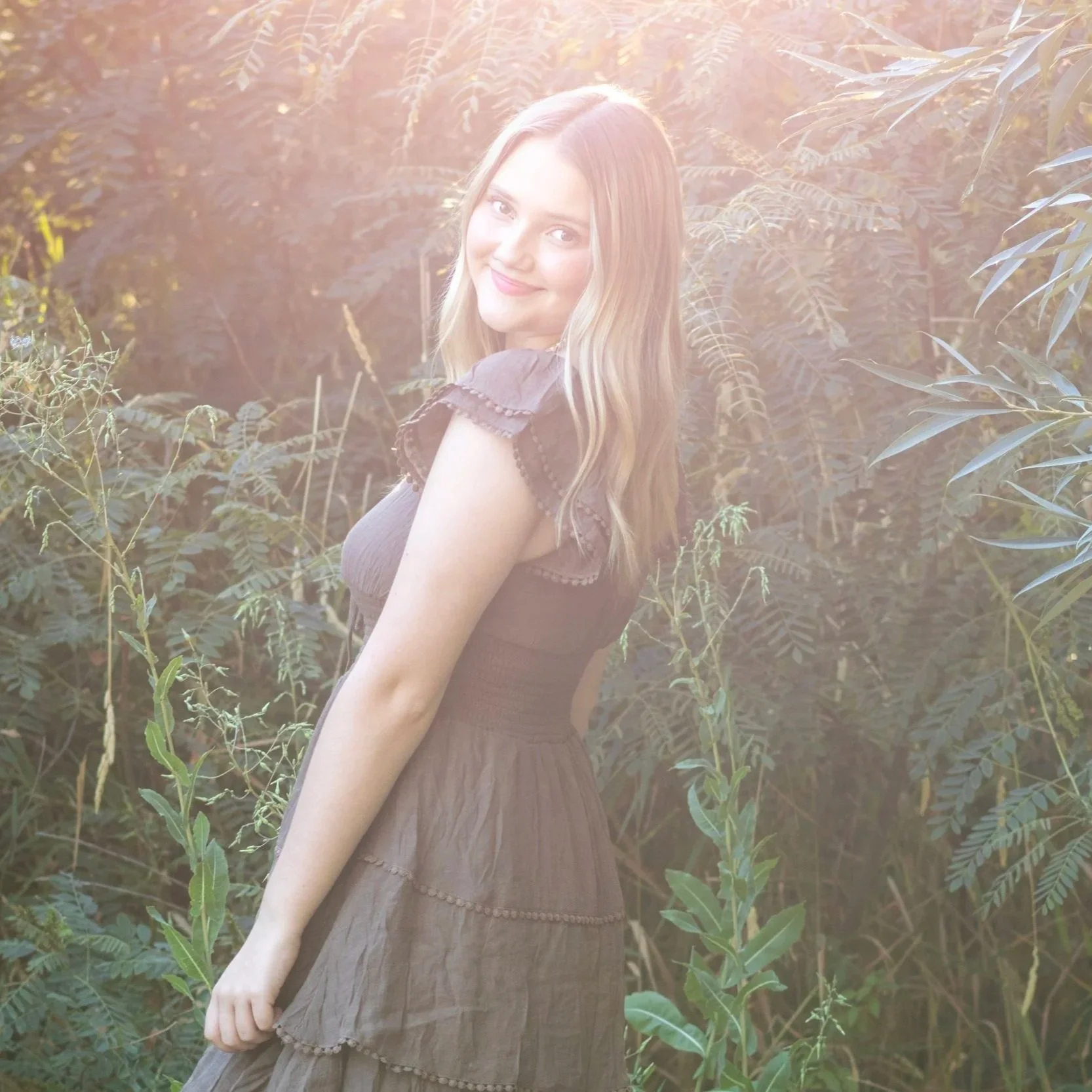 A young woman with long blonde hair smiling in a brown dress outdoors with greenery in the background, sunlight shining from behind. This is from her senior photo session.