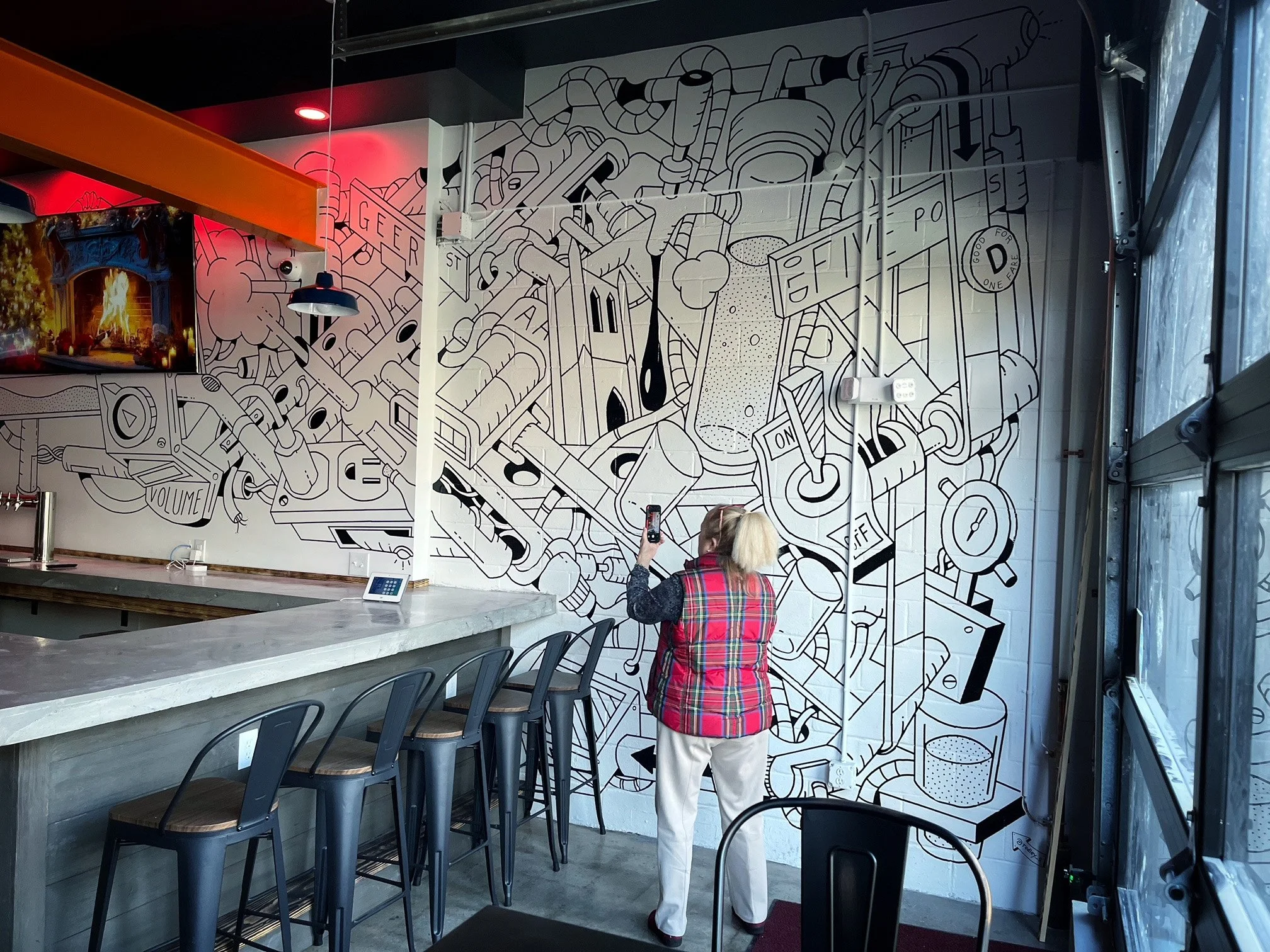 A woman taking a photo of a large black and white mural on a wall inside a room, with a marble counter and bar stools in the foreground.