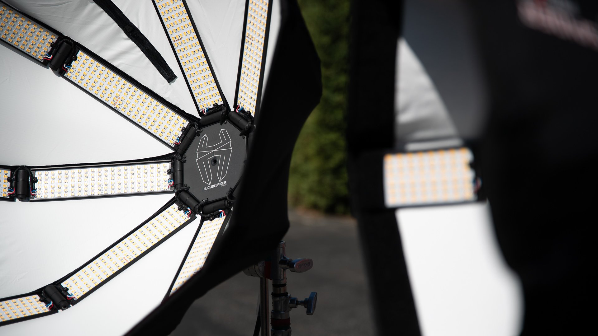 Close-up view of an LED light array with a logo that says 'Hudson Spider' on a black central piece, with additional mounted lights and equipment around it, outdoors.