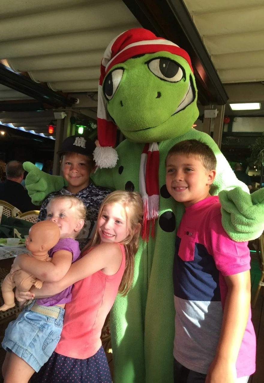 Four children posing with a large green animated character wearing a Santa hat and holiday scarf at a family-friendly venue.