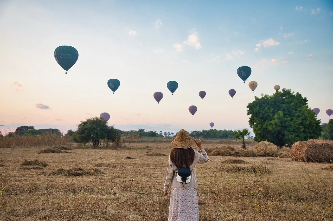 Une femme portant un chapeau de paille regarde des montgolfières dans le ciel au coucher du soleil, dans un champ avec des arbres et des balles de foin.