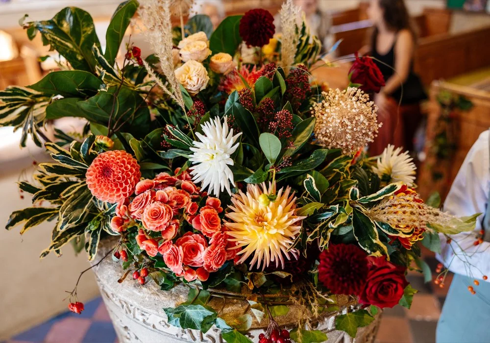 A large floral arrangement in a decorative vase with a variety of colorful flowers including roses, dahlias, and other blooms, along with green leaves and foliage, in a setting resembling a café or restaurant.