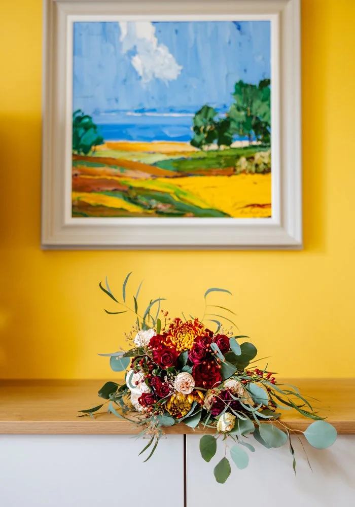 A colorful bouquet of flowers resting on a wooden surface in front of a yellow wall with a landscape painting hung above it.