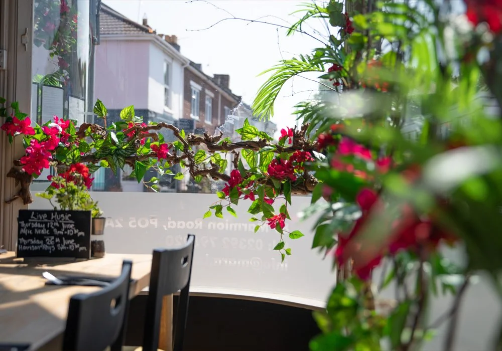 Indoor restaurant or cafe with a window view of residential buildings, decorated with pink and red flowers and green foliage, sunlight streaming in.