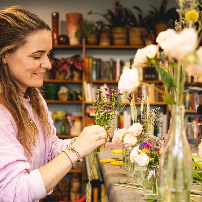 A woman arranging flowers in a florist shop with shelves of books and plants in the background.