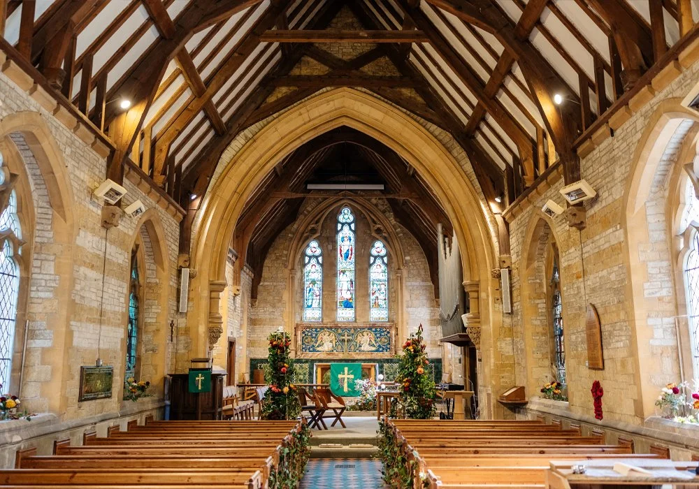 Interior of a small church or chapel with wooden ceiling beams, stone walls, stained glass windows, pews decorated with flowers, and an altar with a green cloth and cross.