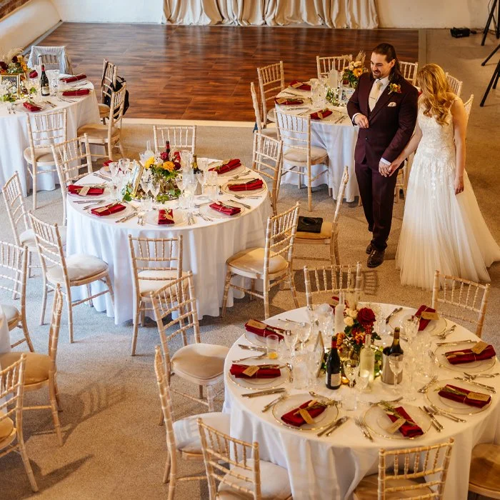 Wedding reception with round tables decorated with white tablecloths, red napkins, and floral centerpieces. Bride and groom walking together.